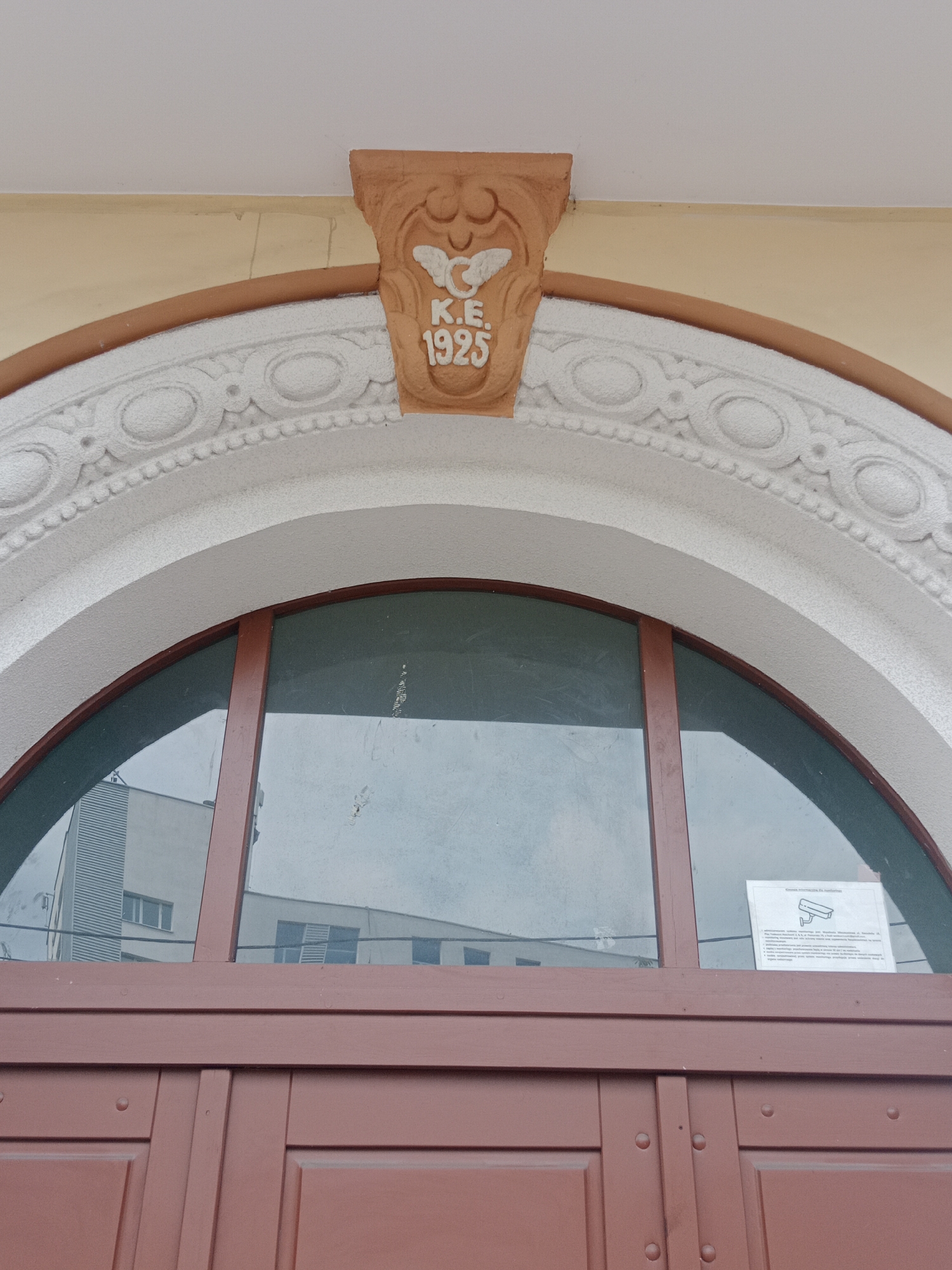 The image shows the upper part of a building's facade in Bydgoszcz, Poland, specifically above a set of double doors. Above the doors is a semicircular window with a dark frame. The window is framed by an ornate, white architectural element with carved circular and linear designs. Above this, a terra cotta decorative piece features the letters "K.E." and the year "1925" embossed beneath a stylized bird or winged symbol. The building facade is painted a pale yellow, and the doors are a reddish-brown color. Reflections of other buildings are visible in the window. A sign, likely a security camera warning, is attached to the right side of the window. The overall scene depicts an architectural detail of a historical building.