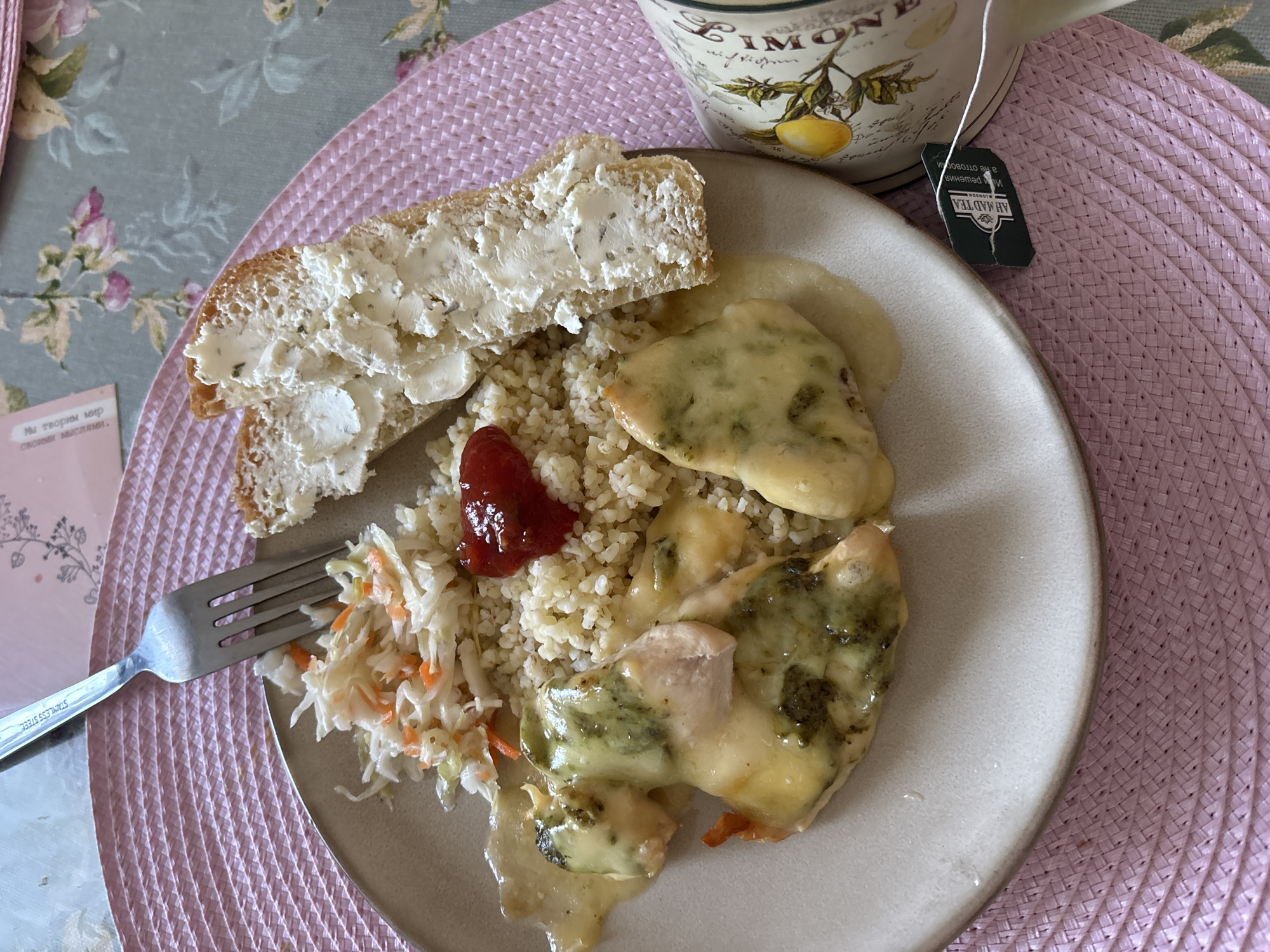 A close-up overhead shot shows a plate of food with chicken, couscous, and coleslaw. The chicken is covered in a creamy, green sauce and cheese. The couscous is topped with a dollop of red sauce. The coleslaw is a mix of shredded cabbage and carrots. To the right of the plate is a slice of bread with butter and a mug of tea. The mug has a lemon design and the tea bag tag reads "AHMAD TEA". The scene is set on a pink placemat with a floral tablecloth in the background. This image was taken in Naberezhnye Chelny, Russia.