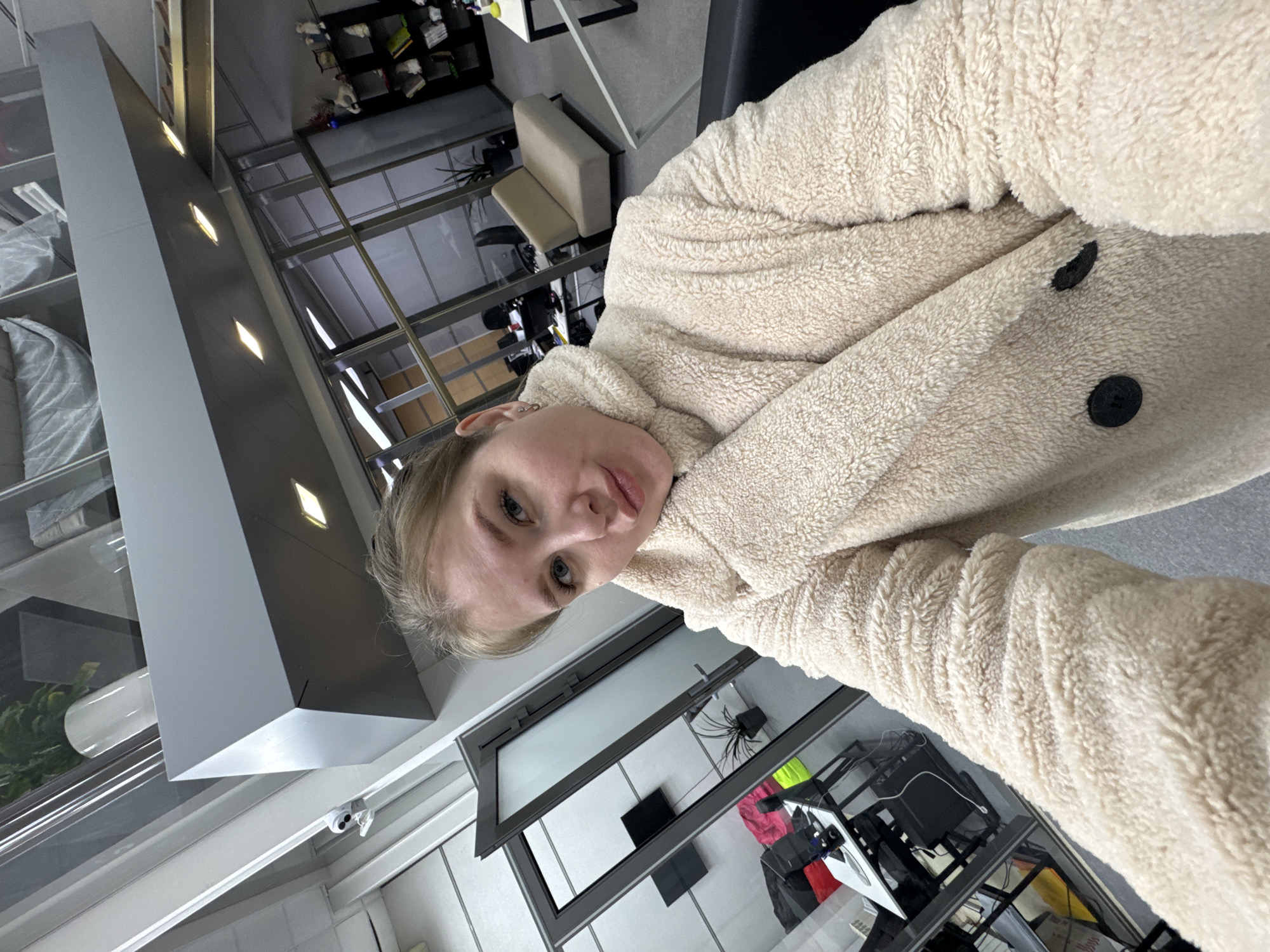 A young woman with light hair and eyes is taking a selfie in an indoor setting. She is wearing a cream-colored, fluffy, possibly fleece or sherpa-style jacket, featuring two visible black buttons on the front. She has multiple piercings on her left ear. Her expression is neutral, and she looks directly at the camera.
The background suggests a modern commercial or office interior. To her left, shelves display neatly organized items, possibly textiles or products. Above these shelves, a grey structural element features several rectangular, recessed light fixtures. A small security camera is mounted on a white ceiling or wall surface.
Further into the background, large glass partitions or windows separate different areas, reflecting portions of the room. Behind these, a light-colored padded bench or chair, a black shelving unit with various items, and a desk or table cluttered with electronics, cables, and bags (including bright pink/red and yellow/green ones) are visible. A potted plant can also be seen behind a glass barrier. The space is well-lit, primarily by artificial sources. The image is oriented sideways, appearing rotated 90 degrees counter-clockwise. No visible text is present. The location context is Perm, Russia.
