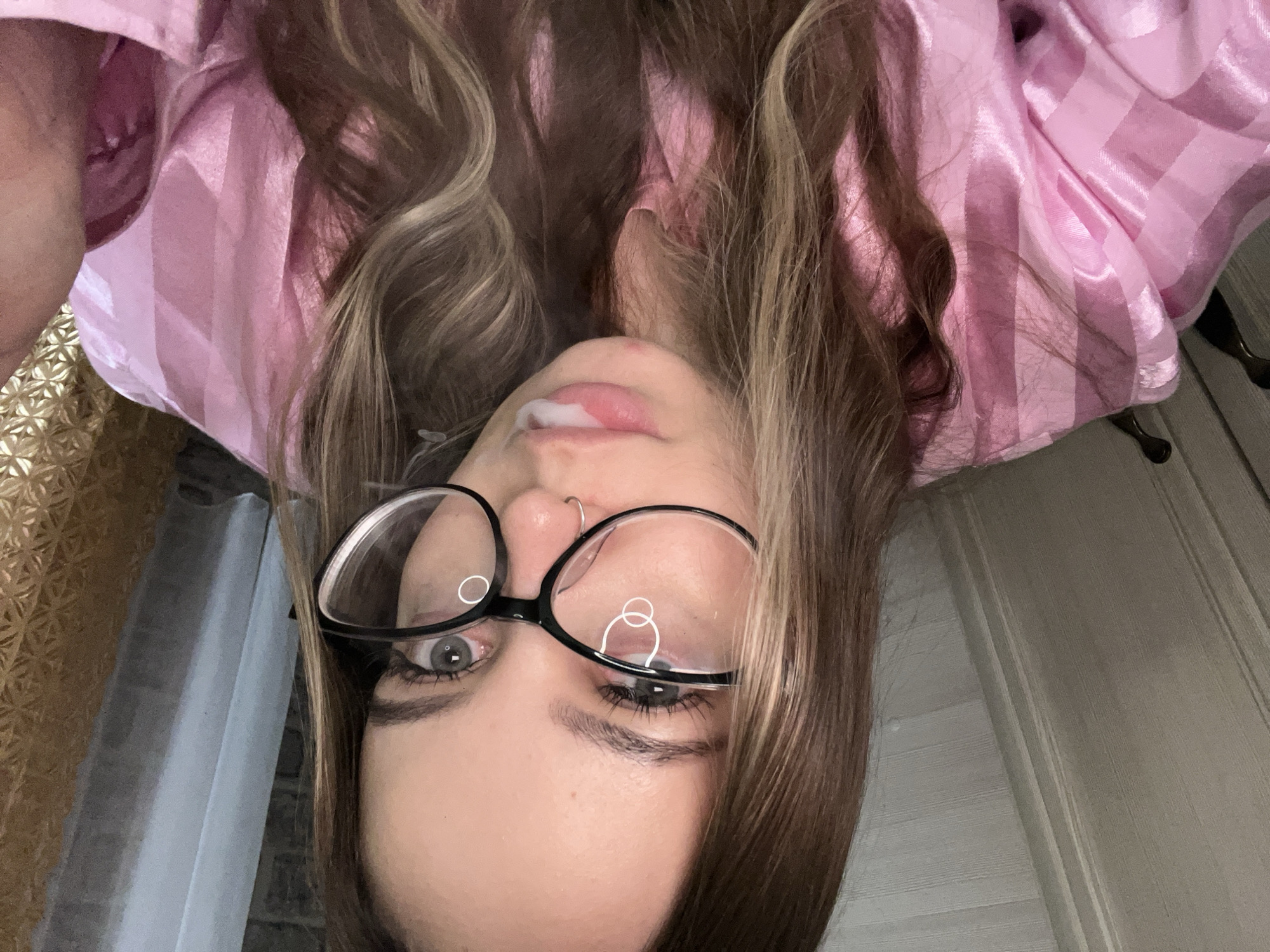 A young woman is seen in an upside-down, close-up selfie. She has long, brown hair with blonde highlights, wearing black-rimmed glasses and a pink and white striped satin robe. She has a nose piercing and is holding saliva on her upper lip. The background appears to be indoors, with a beige patterned curtain visible on the left, and a white wooden door on the right. The city of Novopokrovskaya, Russia is not discernable in this image.