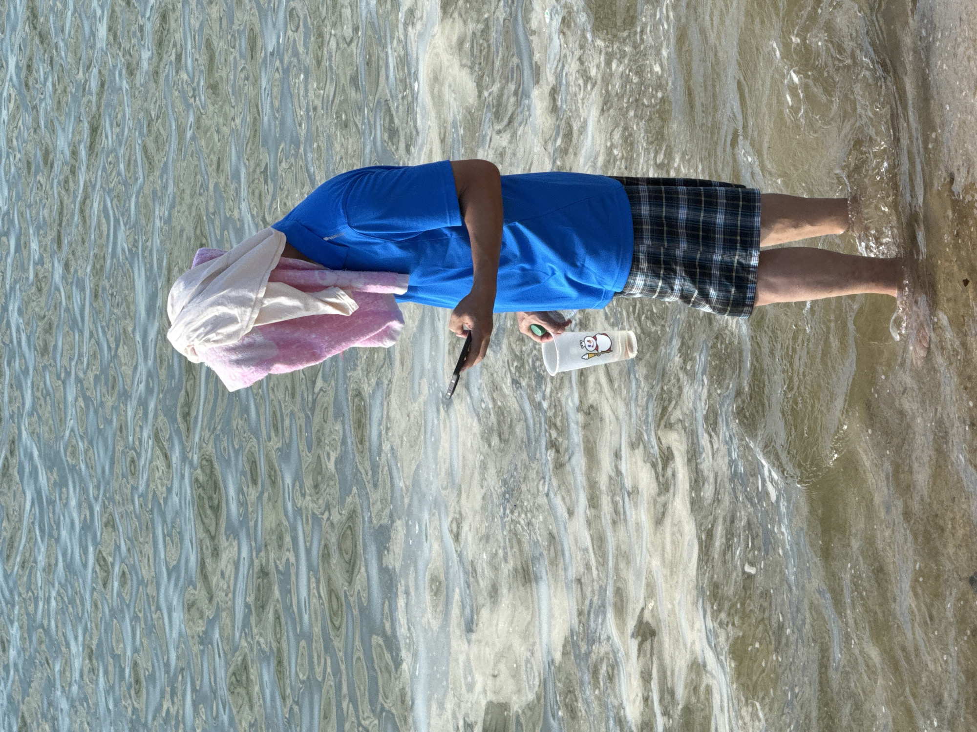 A person stands in shallow water at a beach. The individual is wearing a blue polo shirt, plaid shorts, and has towels draped over their head. They are holding a smartphone in their right hand and a clear plastic cup with a snowman image in their left hand. The water is slightly wavy and appears murky. The background consists of more water and some foam. The scene is shot from a medium distance, with the lower half of the person's body and the water occupying most of the frame. The lighting suggests it is daytime, likely sunny.