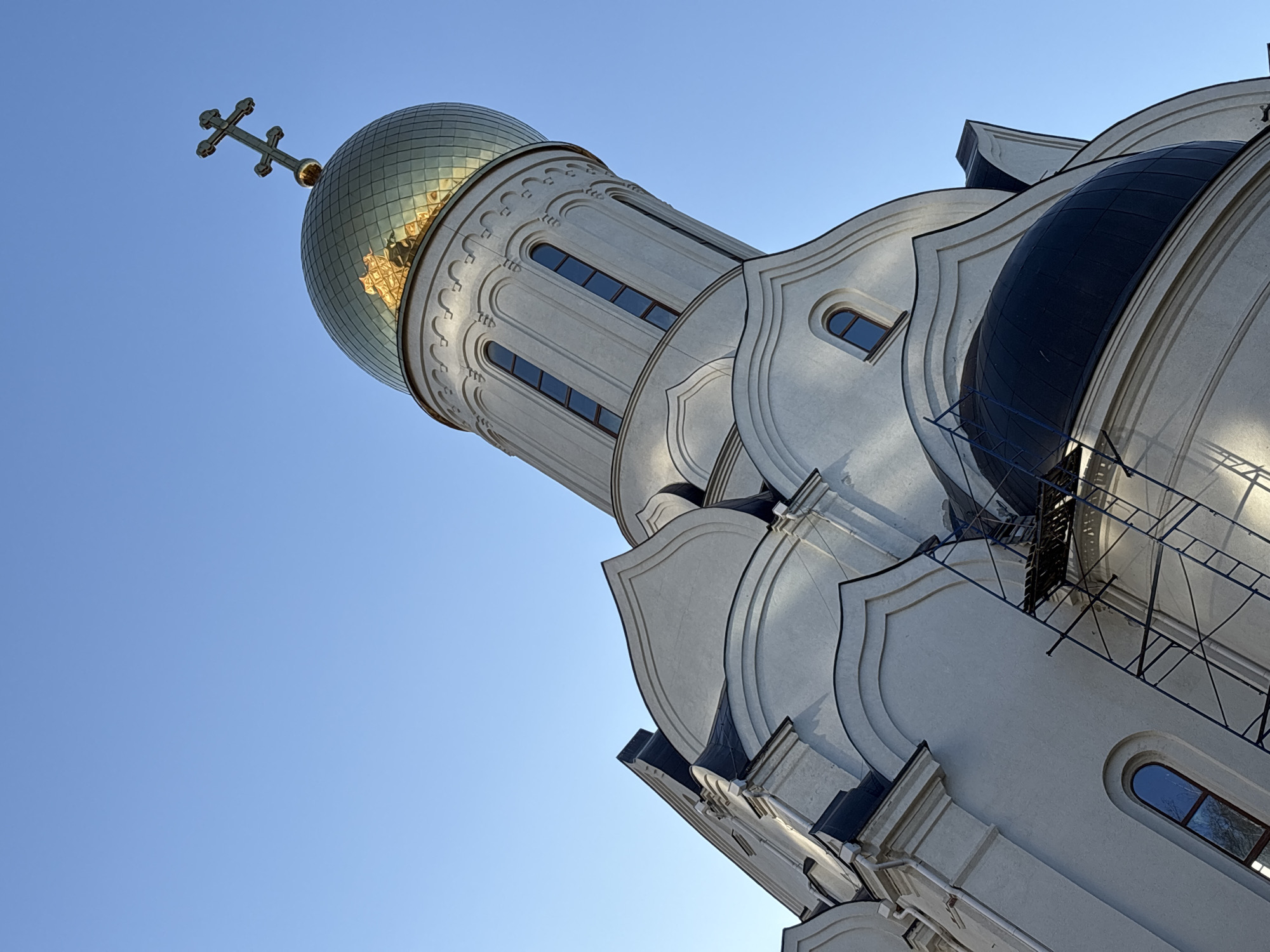 An upward-angled photograph captures the elaborate architecture of an Eastern Orthodox church against a clear blue sky. The most prominent feature is a large, central golden dome, covered in reflective, segmented tiles that shimmer in the sunlight. A traditional Orthodox cross, characterized by its multiple bars, crowns the peak of this dome.
The building itself is constructed from light-colored stone or stucco, appearing off-white or very light grey. It features a bell tower or central structure with several tall, narrow arched windows symmetrically arranged. Decorative elements, including relief patterns and layered, curving facades, adorn the exterior walls. To the right, part of a smaller, darker, possibly black or dark blue dome is visible, contrasting with the lighter main structure. Below this darker dome, some scaffolding or a similar structure can be seen, suggesting ongoing maintenance or construction. Other arched windows, some with dark frames, are scattered across the various sections of the building.
The bright, uniform blue sky indicates a clear, sunny day, likely morning or late afternoon given the directional light that highlights the dome and creates subtle shadows on the building's contours. There are no people visible, and no discernible text. The architectural style, particularly the domes and cross, strongly points to an Orthodox church, consistent with the provided context of Kudrovo, Russia.