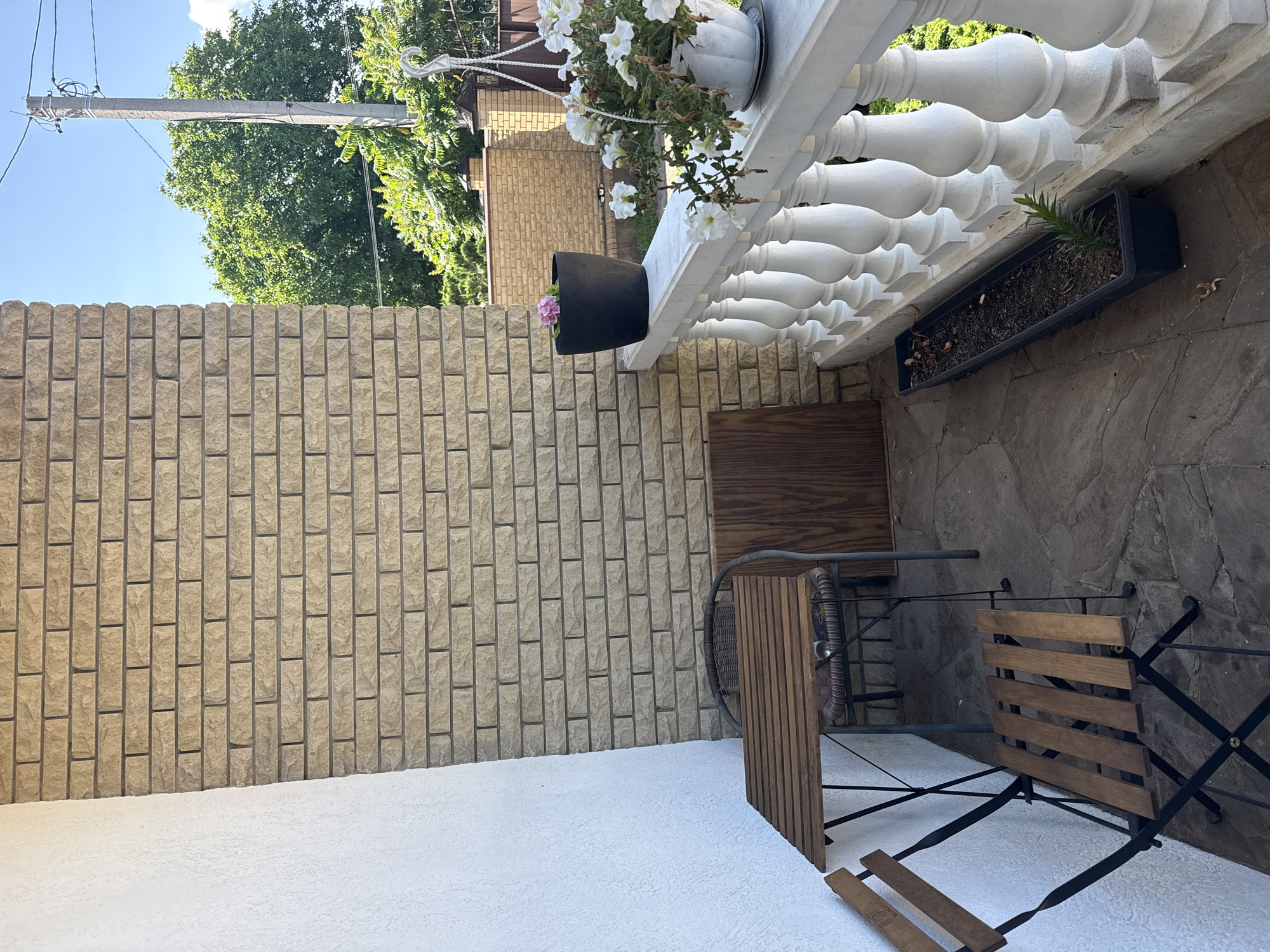 The image depicts a small outdoor seating area on a balcony or patio in Horbanivka, Ukraine. The area is furnished with a small, slatted wooden table and two matching folding chairs, one of which is partially obscured by the table. A dark-colored planter with a single pink flower sits on the railing, and a white planter filled with white petunias hangs from the railing. A long, dark planter box containing soil and a small green plant is also positioned along the railing. The background reveals a beige brick wall and a white textured wall, with trees and a blue sky visible beyond the railing. The ground is paved with stone tiles.