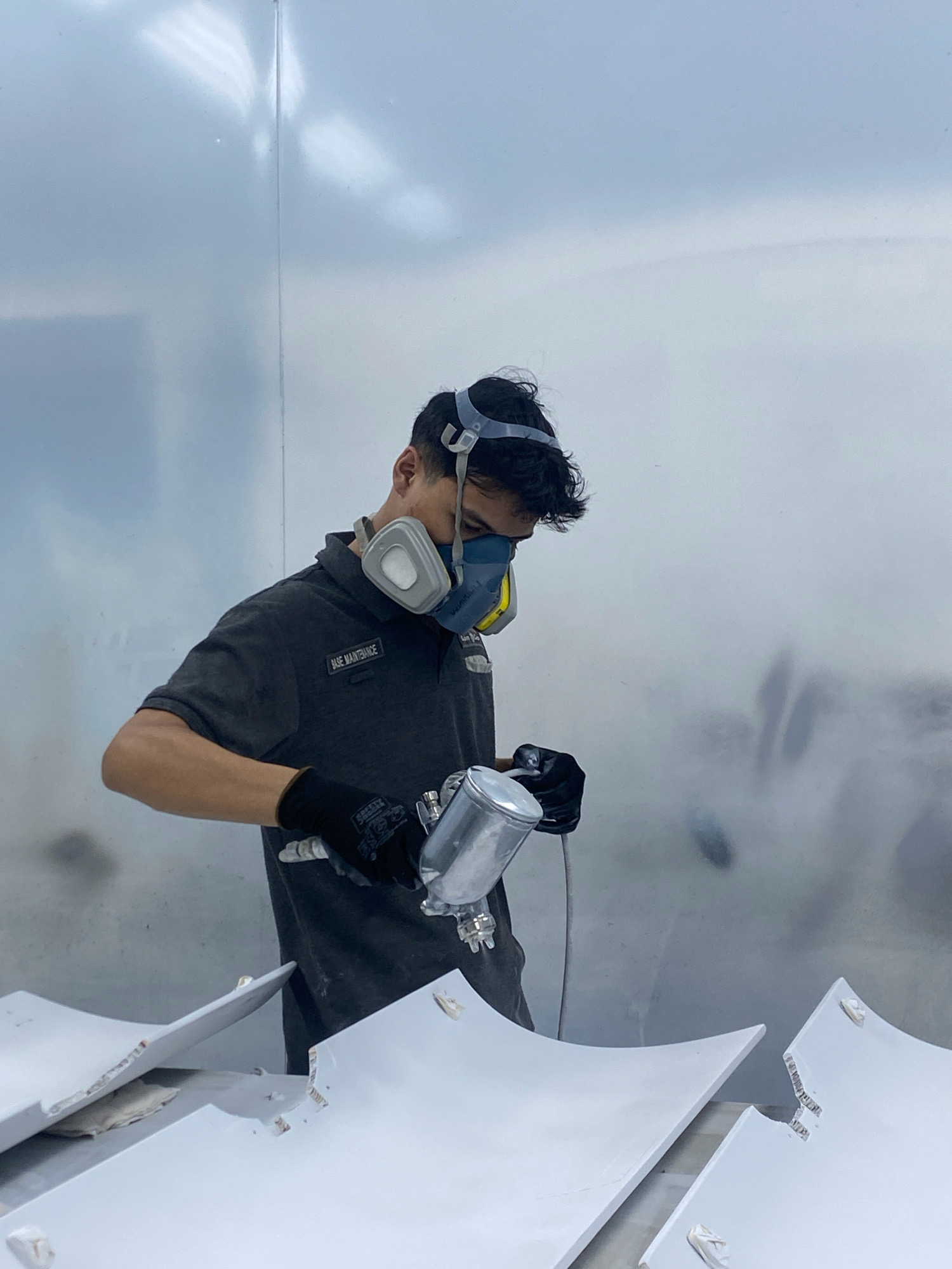 A man is spray painting white panels in a room. He is wearing a gray polo shirt with a patch that reads "BASE MAINTENANCE," black gloves, and a respirator mask. He is holding a spray gun in his right hand. The panels are arranged on a table in front of him. The walls of the room are white and appear to be reflective. This scene is located in a spray booth at a maintenance facility in Batam, Indonesia.