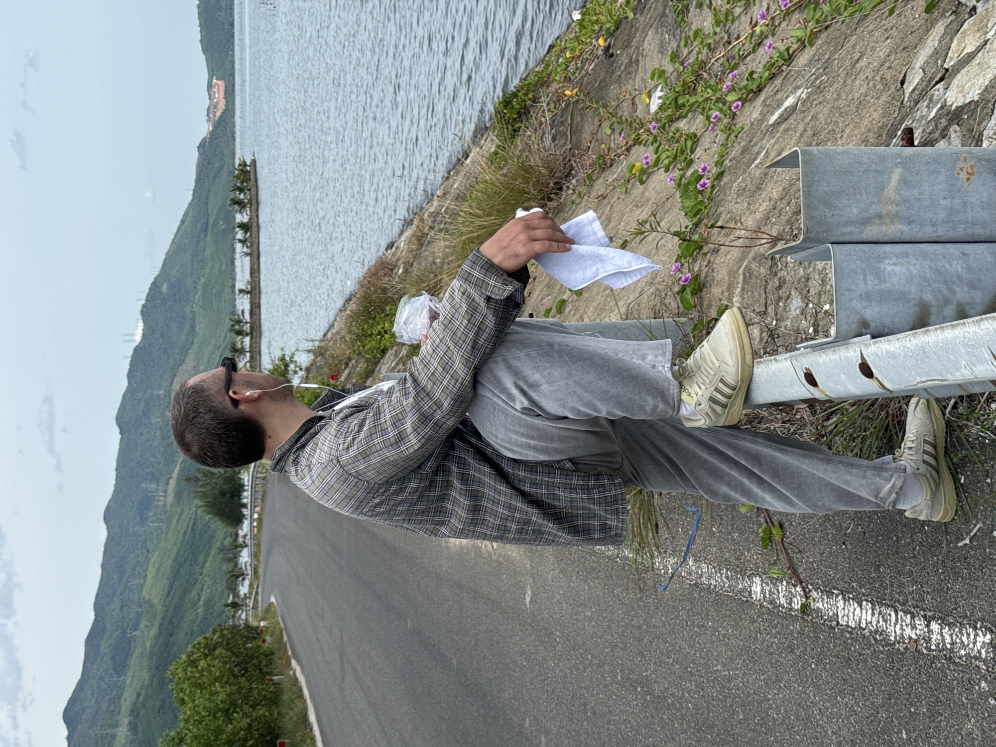 A man stands on the side of a road next to a body of water, with his right leg bent and resting on a guardrail. He is wearing a plaid shirt, grey pants, and light-colored sneakers, and has earbuds in his ears. He is holding a white cloth in his right hand. The road curves to the left, bordered by a grassy embankment. Beyond the water, a lush, green mountain range dominates the background, with a few palm trees visible on a small peninsula in the distance. The sky is overcast. The scene appears to be located near the Thuan An estuary in the city of Huế, Vietnam, known for its coastal roads and proximity to the sea.