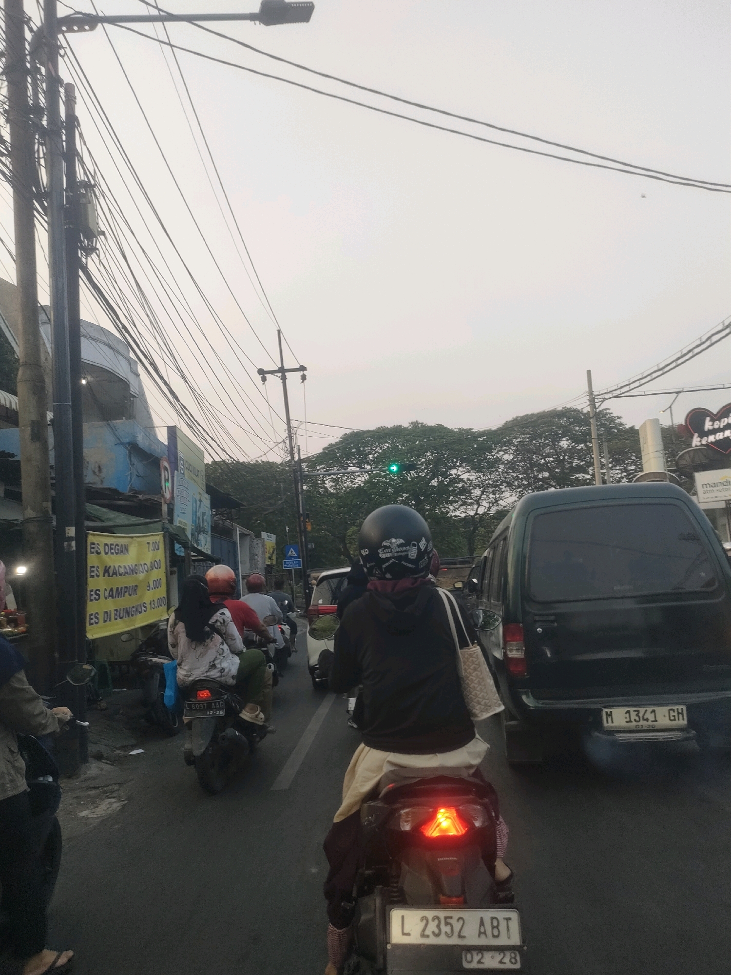 The image depicts a street scene in Surabaya, Indonesia, characterized by moderate traffic congestion and an overcast sky. The foreground is dominated by the rear view of several motorcycles and a dark-colored van. One motorcyclist in the foreground wears a black helmet with white graphics and rides a dark scooter with a visible license plate "L 2352 ABT". The rear brake light of this scooter is illuminated. Another scooter behind it has a license plate "L 6097 AAG". A black van in the middle ground displays the license plate "M 1341 GH".
On the left side of the street, a building with blue and white signage is visible, displaying text that appears to be a price list for drinks like "ES DEGAN" and "ES KACANG HIJO". A lamppost with a multitude of overhead wires is prominent on the left. In the background, a dense canopy of trees forms a verdant backdrop, and traffic lights are visible, with one showing a green signal. The overall atmosphere suggests a typical urban commute in the city.