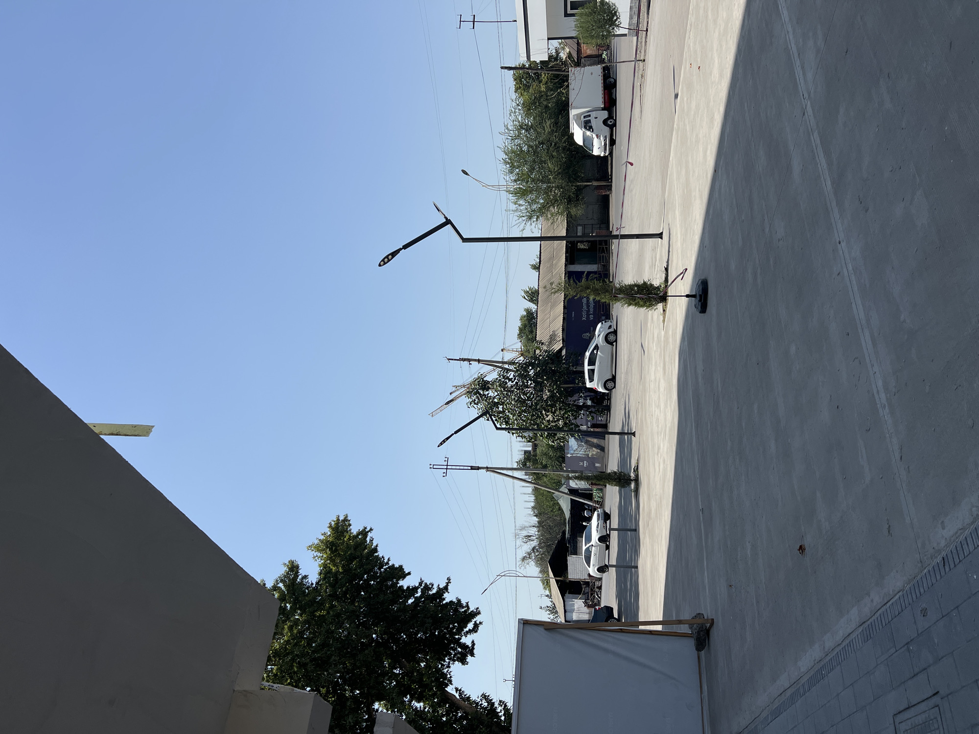 The image depicts an outdoor scene with a clear blue sky. In the foreground, a concrete paved area dominates the lower half of the frame, with some paving stones and a drainage grate visible on the left. A large, light-colored, rectangular structure, possibly a temporary wall or screen, is positioned on the far left.
Further into the scene, several cars are present. A white sedan is parked on the left, and another white sedan is driving on the right. A white truck with a closed cargo box is also visible on the right side. Several streetlights with modern designs are scattered throughout the area.
In the background, there are buildings with corrugated metal roofs, some with awnings. Trees, both deciduous and coniferous, are interspersed among the structures and along the street. Power lines and utility poles are visible against the sky, indicating an urban or semi-urban environment. Construction cranes are also present in the far background, suggesting ongoing development. The overall scene appears to be a public space or a commercial area, likely in a city.