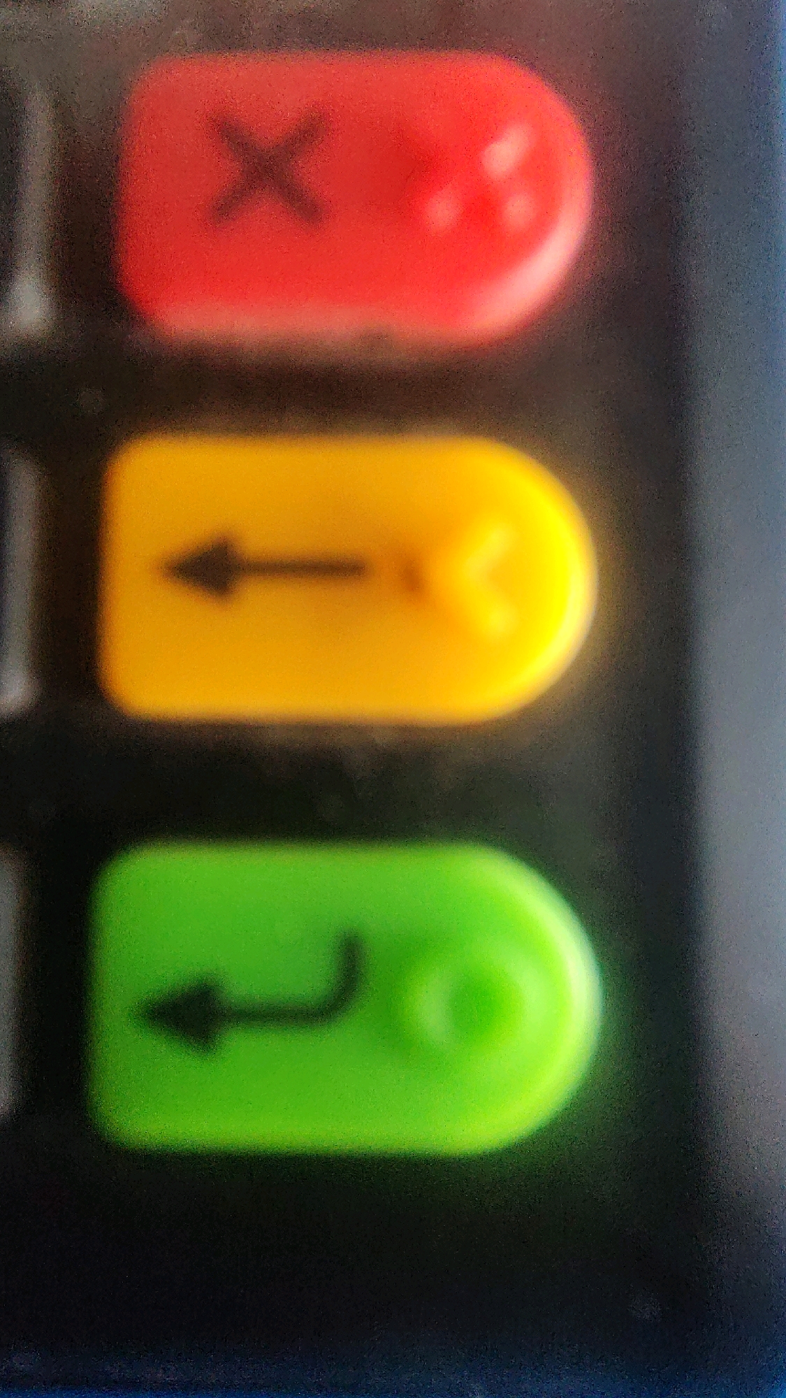 The image displays a close-up, slightly out-of-focus view of three buttons on a device. The top button is red with a black "X" symbol. Below it, a yellow button features a black leftward-pointing arrow. The bottom button is green and shows a black symbol that resembles a leftward-pointing arrow with a curved tail, commonly known as an "enter" or "return" key. The buttons appear to be part of an electronic device's keypad, possibly a calculator, point-of-sale terminal, or remote control. The background is dark and textured, suggesting a matte surface. The image is a static, detailed shot of these specific buttons. The location of Perundurai, India, is not discernible from the visual information provided in the image.
