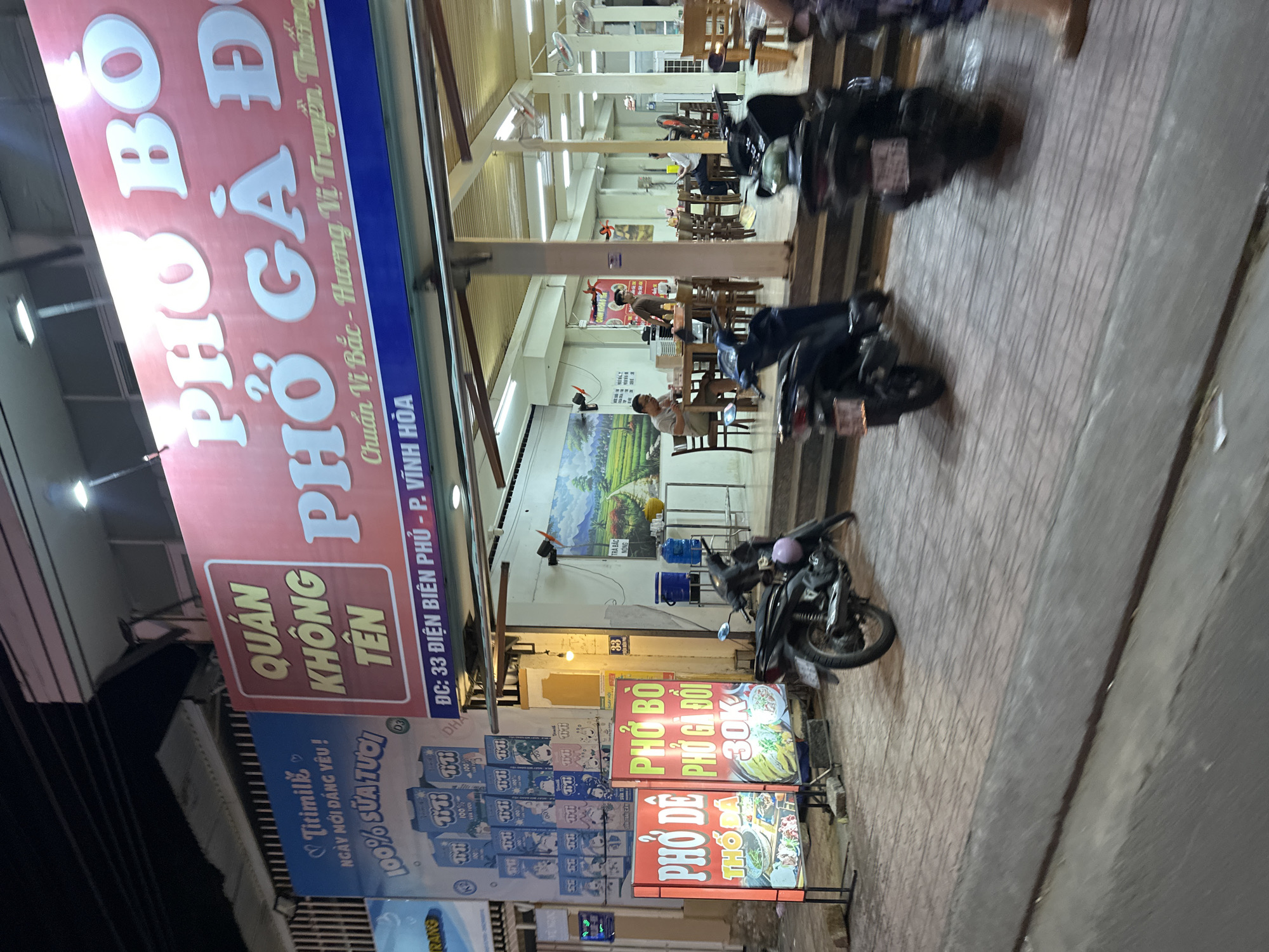 The image shows a street scene in what appears to be the Bac Nha Trang Ward of Nha Trang, Vietnam. The focus is on a restaurant named "Quán Không Tên" (Restaurant Without a Name), which serves Pho Bo (Beef Noodle Soup) and Pho Ga (Chicken Noodle Soup). The large red sign overhead prominently displays the restaurant name and the types of pho offered. Smaller signs on the sidewalk advertise Pho Bo, Pho Ga Doi, and Pho De (Goat Noodle Soup) at 30K. A sign for Titimilk is also visible. Several motorbikes are parked along the sidewalk, suggesting they belong to customers. The restaurant's interior is illuminated, showing tables, chairs, and customers inside. The street is tiled.