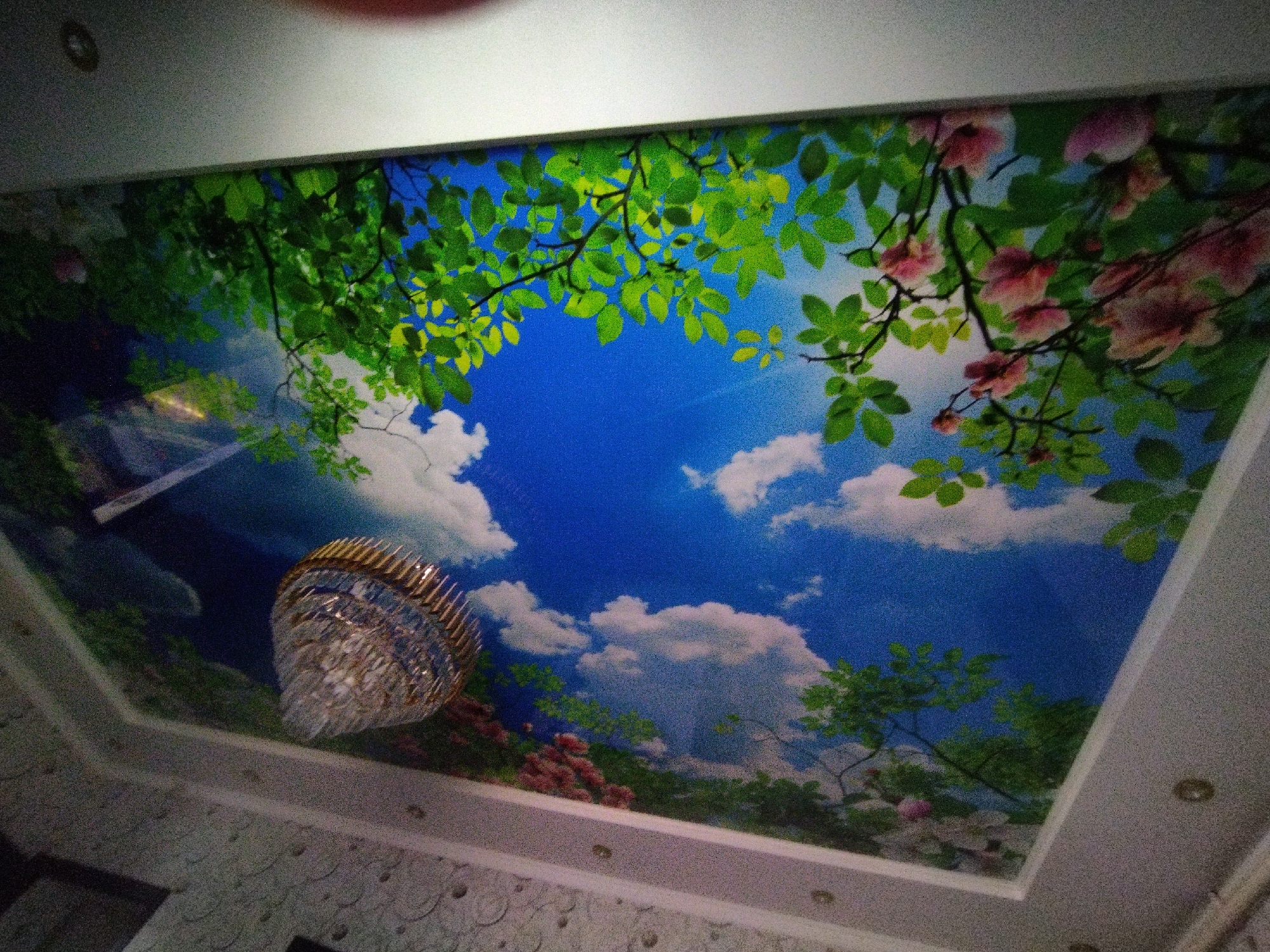 The image shows a rectangular ceiling mural depicting a bright blue sky with scattered white clouds and sunlight filtering through lush green leaves and pink flowers. A modern crystal chandelier hangs from the center of the ceiling. The ceiling is recessed, with a white border. The mural creates an illusion of an open sky. The location is not specified in the image and cannot be determined to be in Мраморное, Kyrgyzstan.