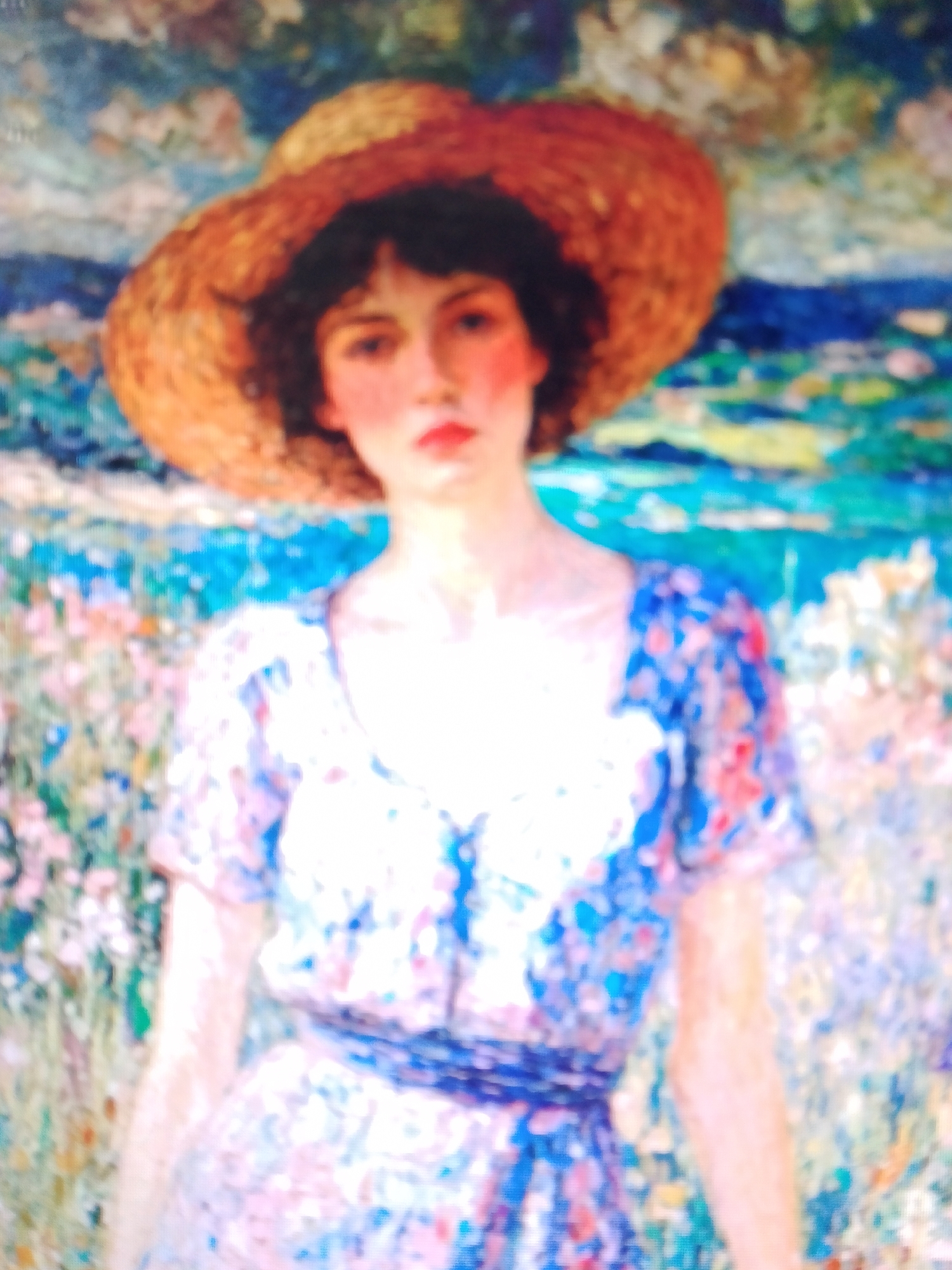 A painting depicts a young woman wearing a wide-brimmed straw hat and a floral dress. Her face is turned slightly to the viewer, with her lips parted. She stands in a field of flowers with a blurred background of greenery and water. The style of the painting is Impressionistic, with visible brushstrokes and a soft, dreamlike quality. The woman's skin is fair, and her hair is dark. She is wearing a blue and white floral dress with a ruffled neckline. The hat is made of straw and has a wide brim that casts a shadow over her face. The flowers in the field are a mix of pink, white, and yellow. The background is a blur of blue and green, suggesting a landscape with water and trees.