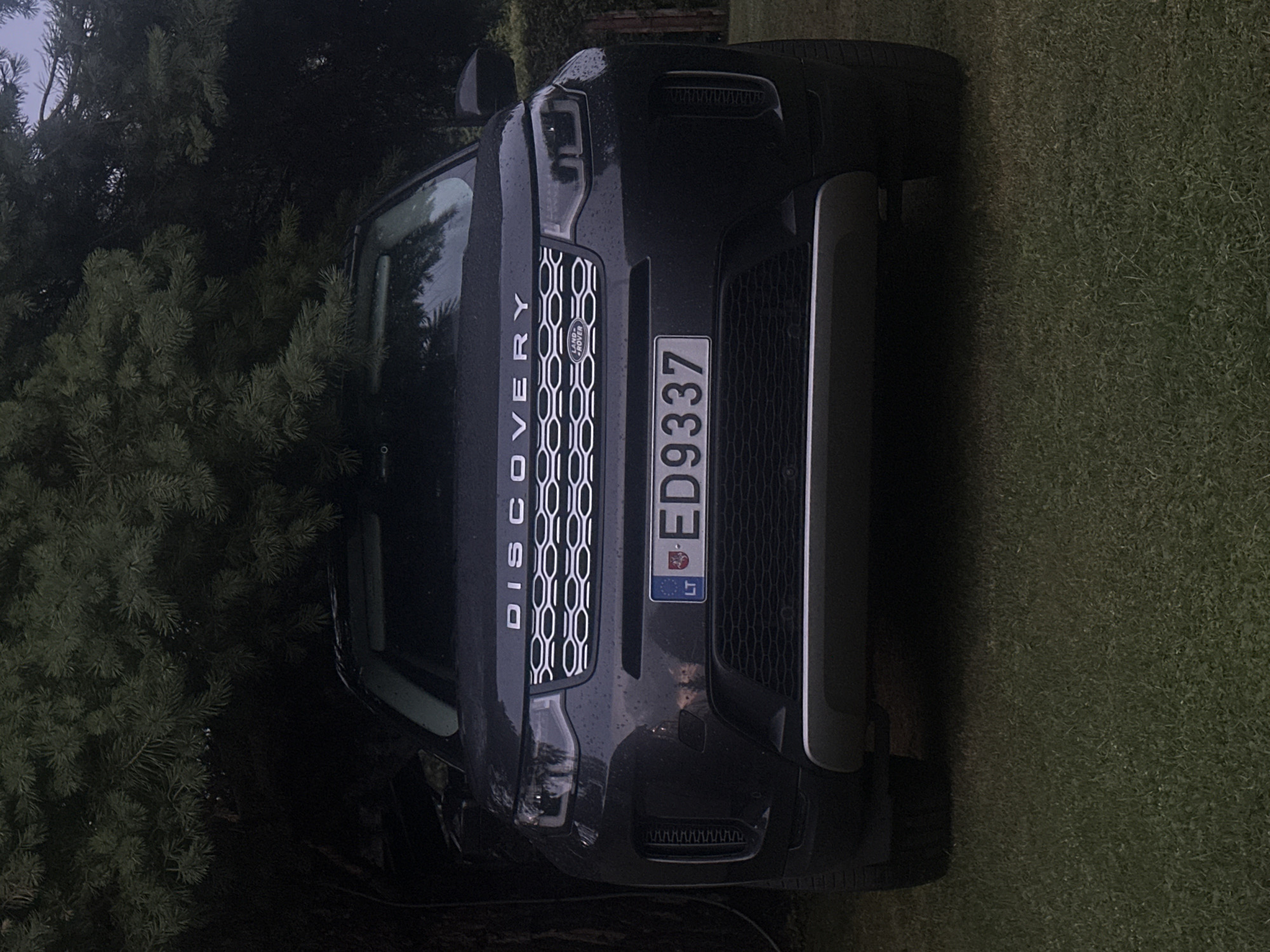 A dark gray Land Rover Discovery Sport is parked on a green lawn, facing the camera. The vehicle is positioned beneath a large, dark green conifer tree, with its branches casting shadows over the car. The front of the car is visible, including the headlights, grille with the "DISCOVERY" emblem and Land Rover logo, and a license plate that reads "ED9337" and has the "LT" country code. The car appears to be in good condition. The background shows more of the conifer tree and hints of a fence and some foliage. The lighting suggests it is either early morning or late evening. No people are visible in the image. The location is identified as Lapės, Lithuania.