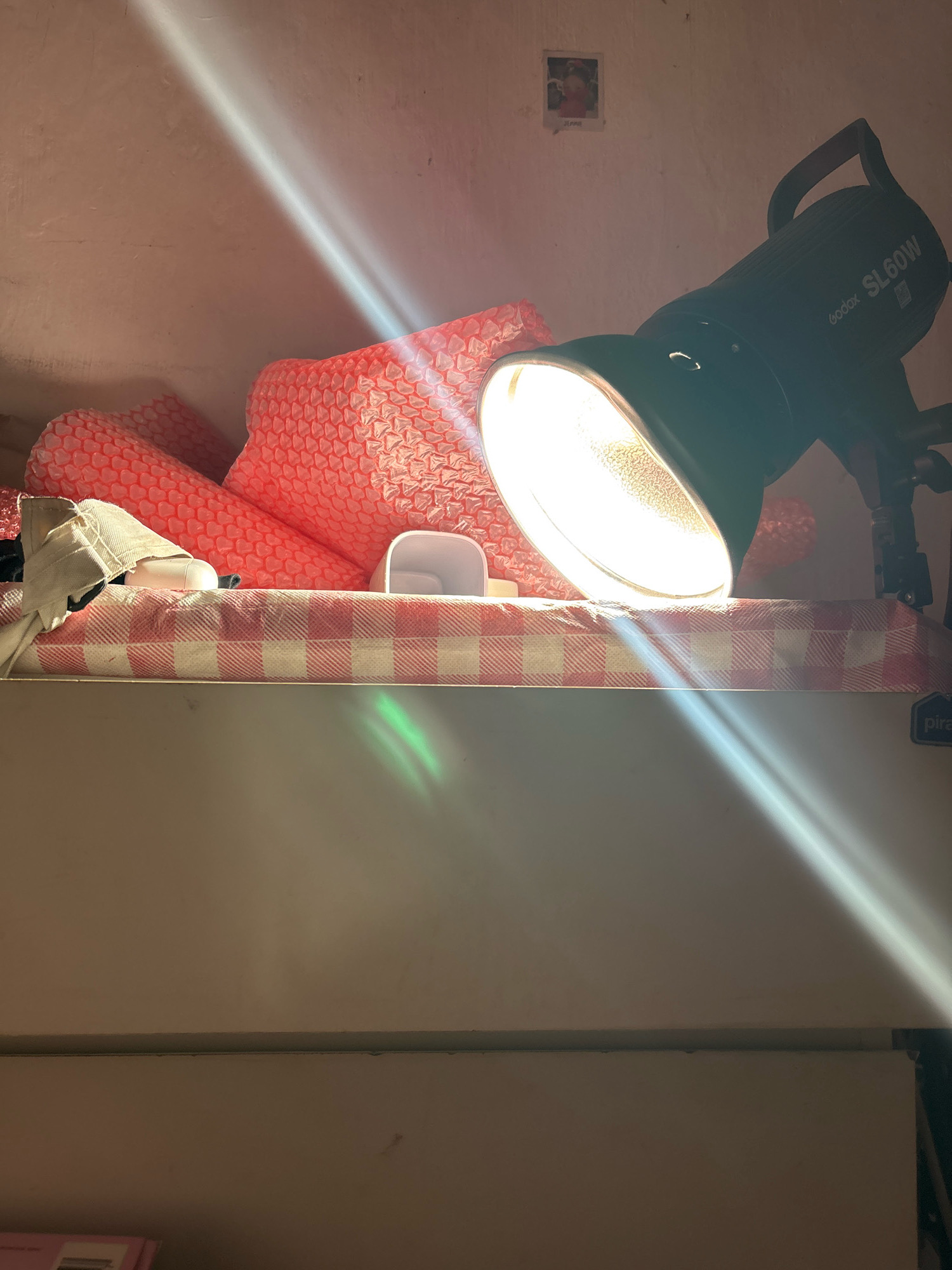 The image depicts an indoor scene, possibly a studio or storage area, with several objects arranged on a surface covered with a red and white checkered cloth. A prominent feature is a black Godox SL60W light fixture with its bright bulb illuminated, casting strong light rays across the scene.
In the background, there are some pink, bubble-wrap-like objects and a small framed picture on the wall. A light-colored fabric object, possibly a mask or cloth, is draped over some of the pink items. A small white object, resembling a container lid, is also visible.
The light fixture is positioned towards the right side of the frame, angled downwards. Light rays from the fixture cut across the image diagonally, creating dramatic highlights and shadows. A faint green reflection is also noticeable on the checkered surface. The overall setting appears somewhat cluttered, with objects piled up.
The provided context suggests this scene is located in North Jakarta, Indonesia. However, the image itself does not contain any visual cues that would specifically indicate this location. The details visible are typical of an indoor domestic or semi-professional setup and do not offer any geographical identifiers.