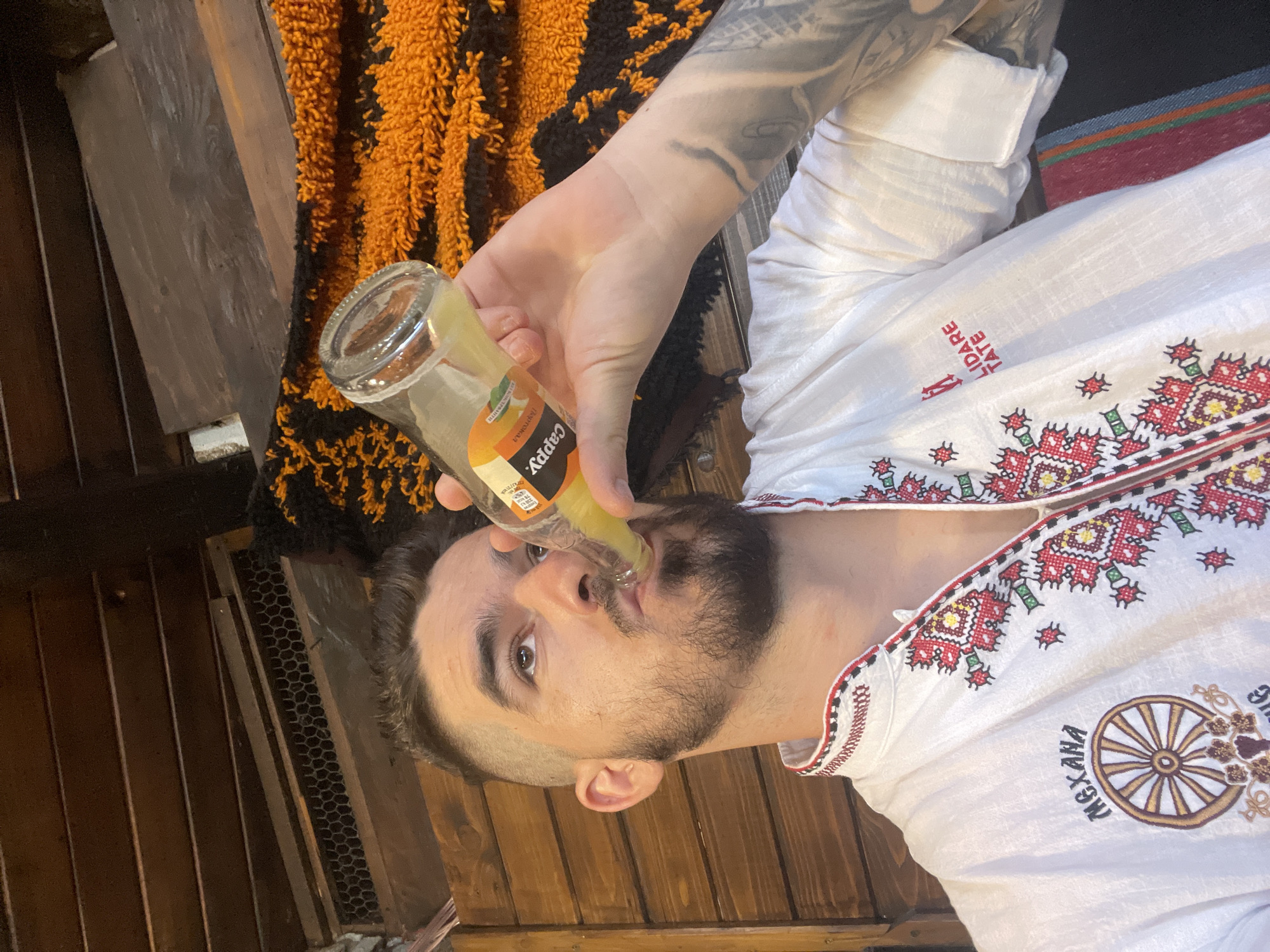 In this close-up, low-angle shot, a man with short, faded hair and a beard drinks from a glass bottle of Cappy juice. He wears a white, long-sleeved shirt with intricate red, white, and green embroidery on the collar and down the front. The shirt also features embroidered text reading "MEXANA" above a wheel design on the left side, and "LIDARE TATE" in red on the right. His right arm, visible on the right side of the frame, is heavily tattooed. The background consists of rustic wooden walls and beams, with a textured orange and black fabric hanging behind him. The man is indoors, possibly in a restaurant or traditional establishment, with a warm, dimly lit ambiance.