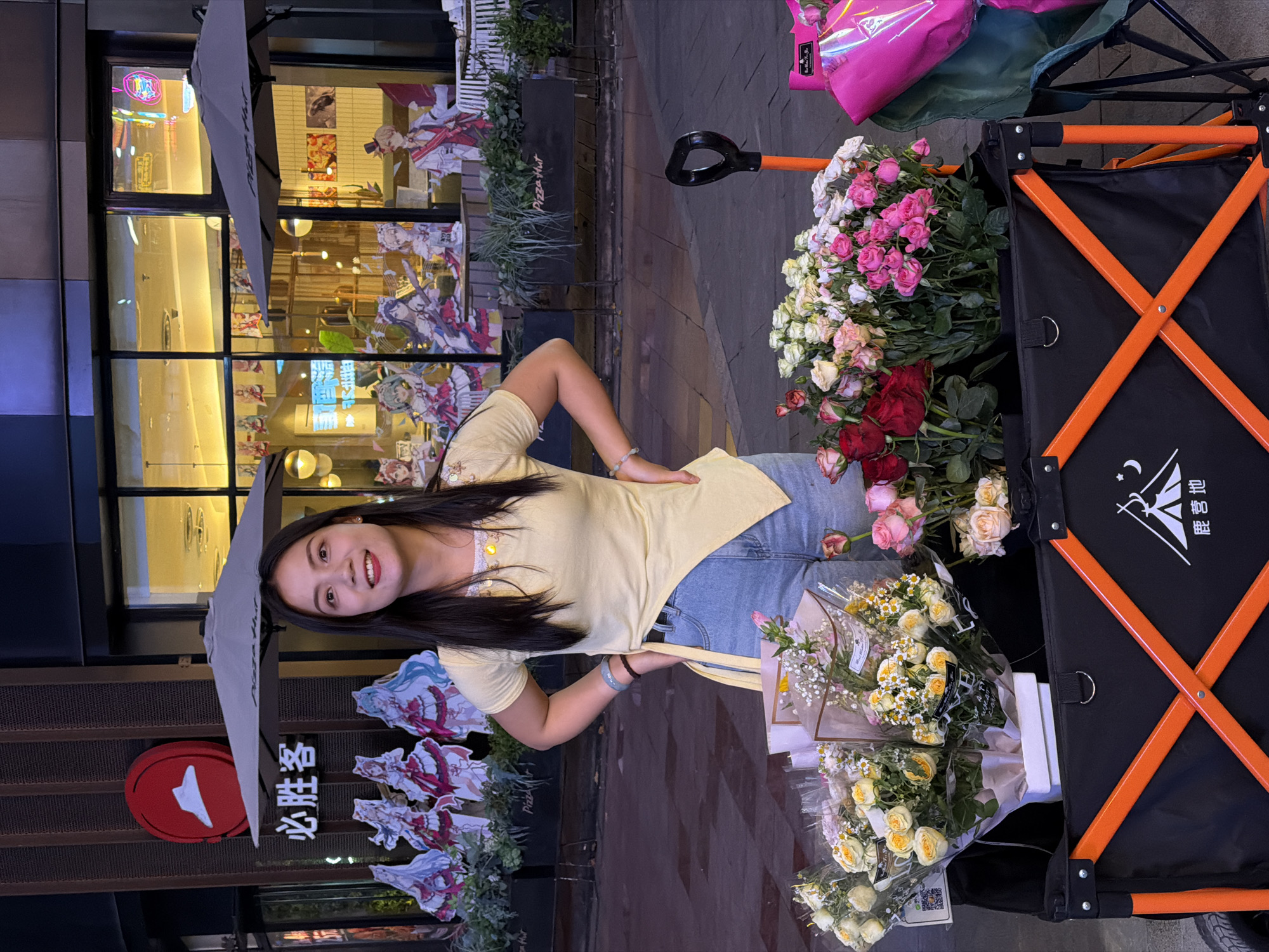 A young woman poses next to a flower cart in Chongqing High-tech Zone, China. The woman is wearing a yellow shirt and jeans. The flower cart is full of various bouquets of flowers. Behind the woman is a Pizza Hut restaurant. The restaurant's windows are decorated with anime posters. An umbrella is visible above the woman. The overall setting is an urban street scene at dusk or night.