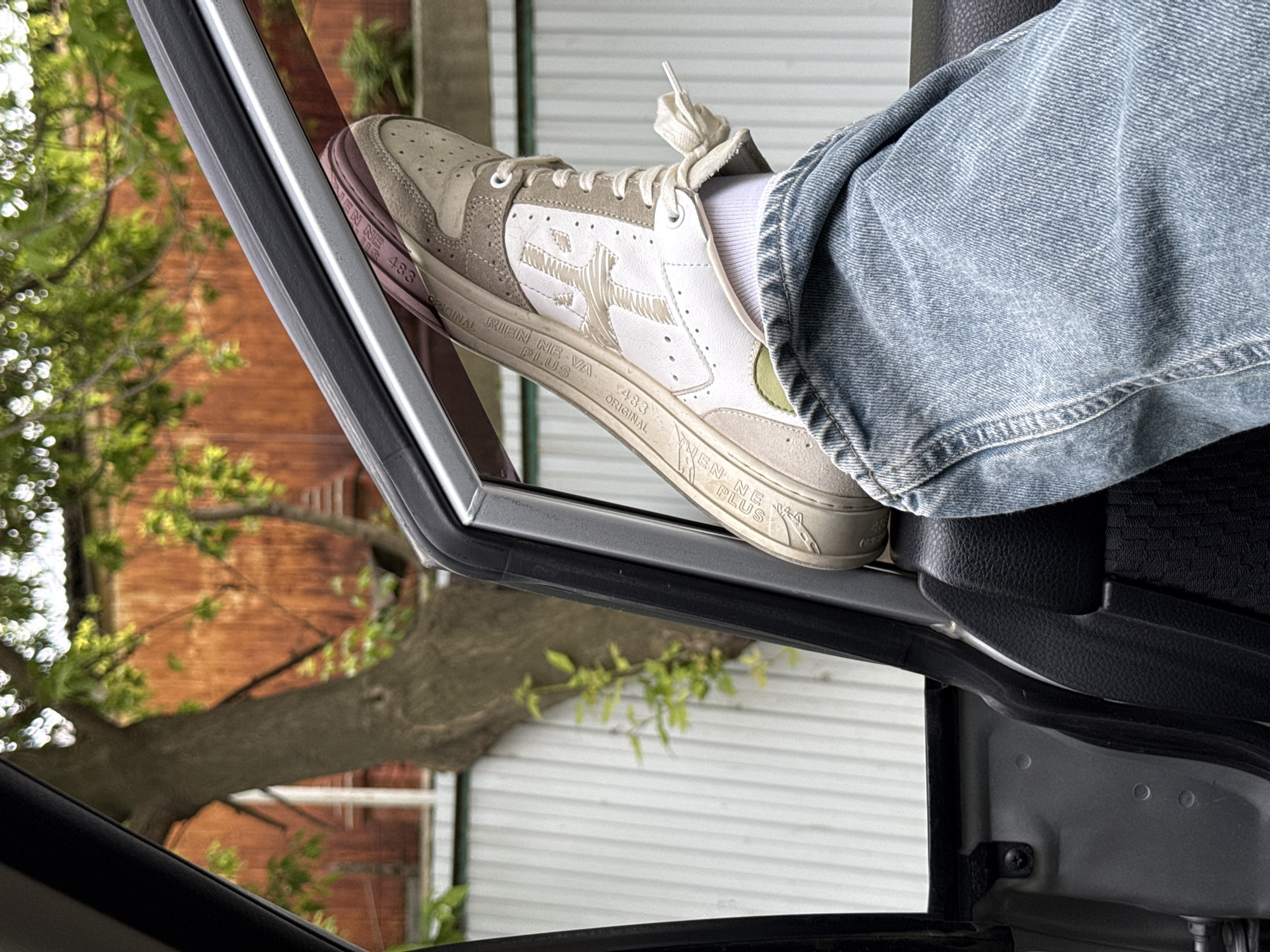 A low-angle shot captures a person's leg, clad in light blue jeans and a white sock, resting a sneaker on the lower frame of an open car door. The sneaker is predominantly white and beige with a perforated toe box, lace-up closure, and a visible green accent on the side. Text "RIEN NE VA PLUS" and "483 ORIGINAL" is visible along the pinkish-brown sole of the shoe, with "483" appearing again on the upper. The car's black door panel and a metallic grey door frame are in the foreground.
Beyond the car, an outdoor scene is visible, inverted, suggesting the photograph was taken with the car door open and the camera angled upwards or the image has been rotated. A large tree trunk with green foliage dominates the midground. In the background, sections of a reddish-brown brick building and a white-sided building are discernible. The bright lighting suggests it is daytime. The scene provides cues of an urban or suburban environment, set in Vladivostok, Russia.