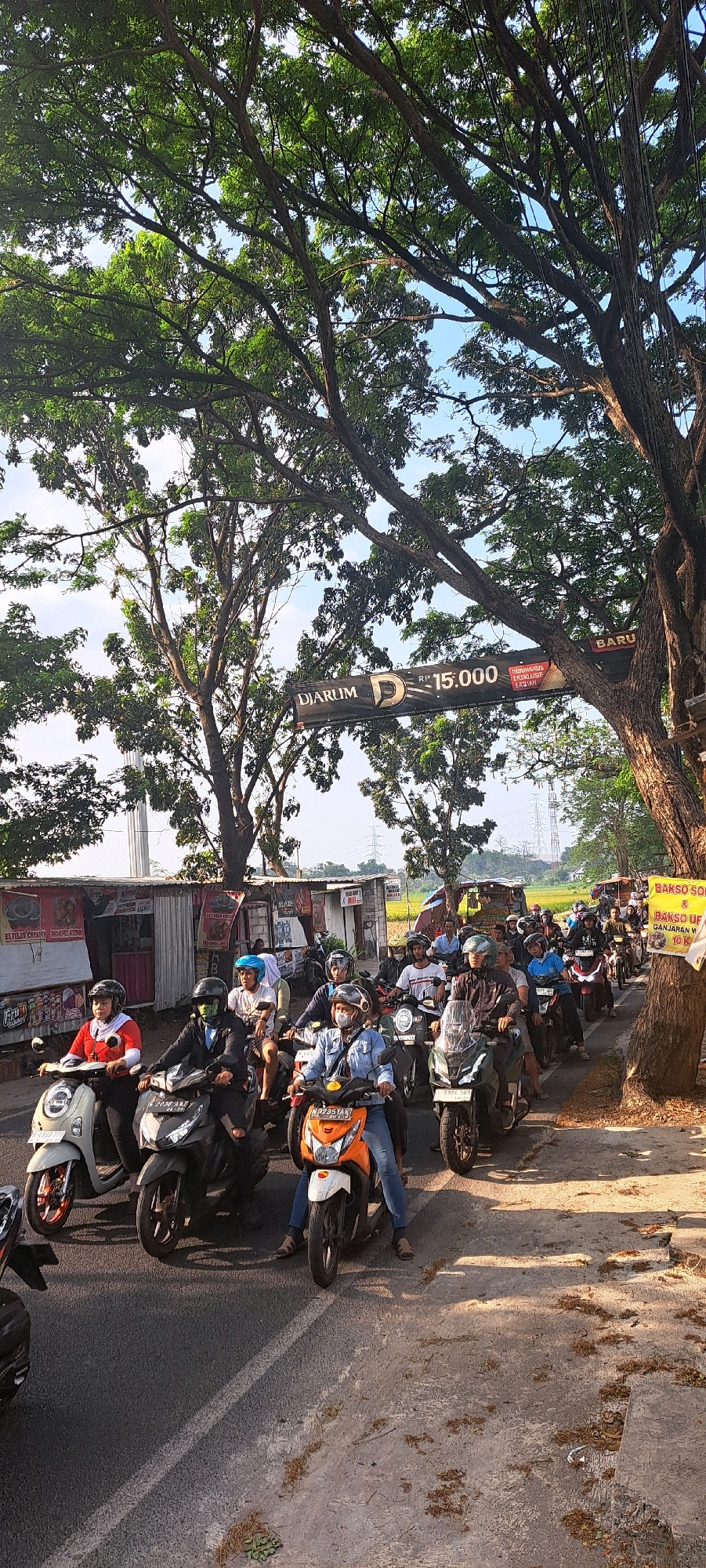 The image depicts a traffic scene in Randupitu, Indonesia, characterized by a line of motorcycles and their riders on a paved road. A large tree with dense foliage overhangs the road, casting shadows. In the background, there are several small roadside stalls or shops, and beyond them, a rural landscape with rice paddies and power lines. A banner is suspended across the road, displaying "DIARUM" and "RP 15.000". The riders, mostly men, are wearing helmets and casual clothing. Some are looking ahead, while others are interacting with each other. The atmosphere appears to be one of everyday traffic flow in a semi-urban or rural Indonesian setting.