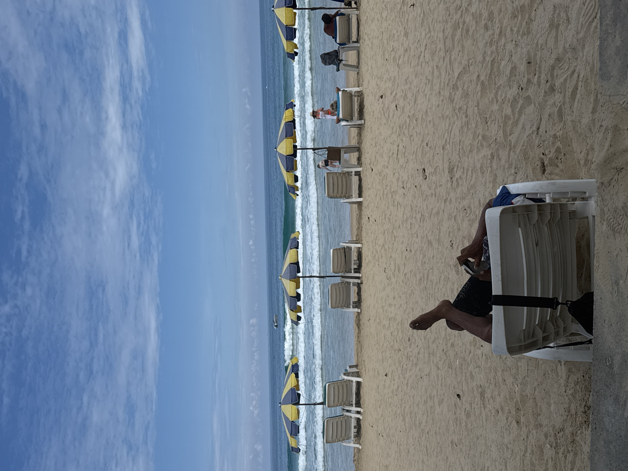 A medium shot captures a beach scene with a person relaxing on a lounge chair in the foreground. The person, seen from the waist down, is lying on their back with their legs extended and crossed at the ankles. They are wearing dark shorts and appear to be holding a phone or similar device.
In the midground, several empty lounge chairs are arranged in a row on the sand, each accompanied by a beach umbrella with alternating yellow and blue panels. The umbrellas are open, providing shade. Beyond the chairs and umbrellas, gentle waves roll onto the sandy shore. The ocean stretches to the horizon under a partly cloudy blue sky. A small boat can be seen in the distance on the water.
Further down the beach, other beachgoers are visible, including a woman in an orange bikini lying on a lounge chair and a child standing near an umbrella. Two men are also seated on lounge chairs, with one appearing to be reading.
The overall environment is a sunny beach with a calm sea and a few scattered clouds. The perspective is from a slightly elevated position, looking towards the ocean.