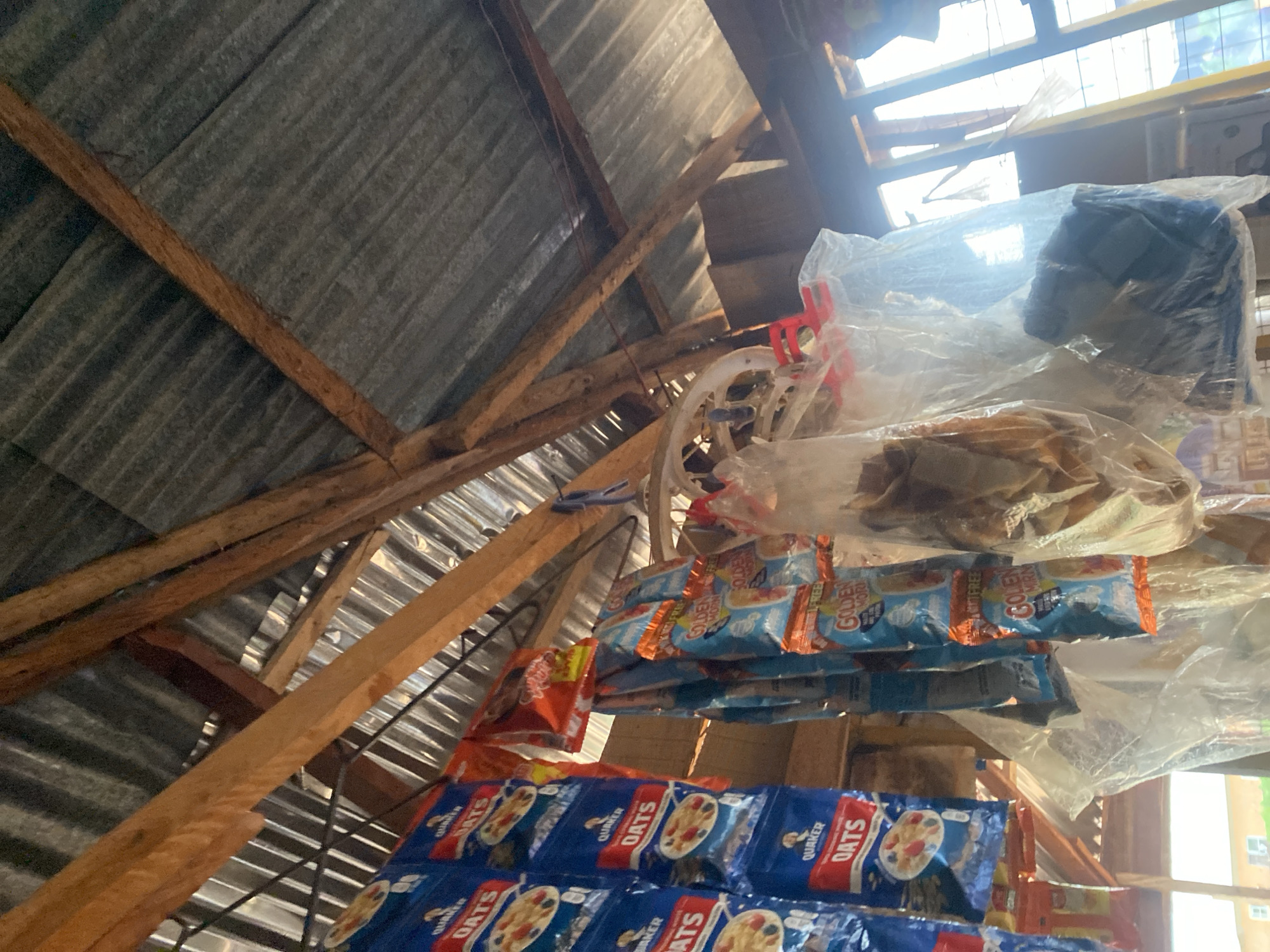 This image depicts an outdoor stall in Lafia, Nigeria, sheltered by a corrugated metal roof supported by wooden beams. The stall displays various food items, including Quaker Oats cereal boxes and "Golden Morn" cereal packets, hanging from a makeshift rack. Some items are packaged in clear plastic bags, one of which contains what appears to be dried fish. The background reveals windows with natural light streaming through, illuminating the scene. The overall impression is of a small, informal market setting.