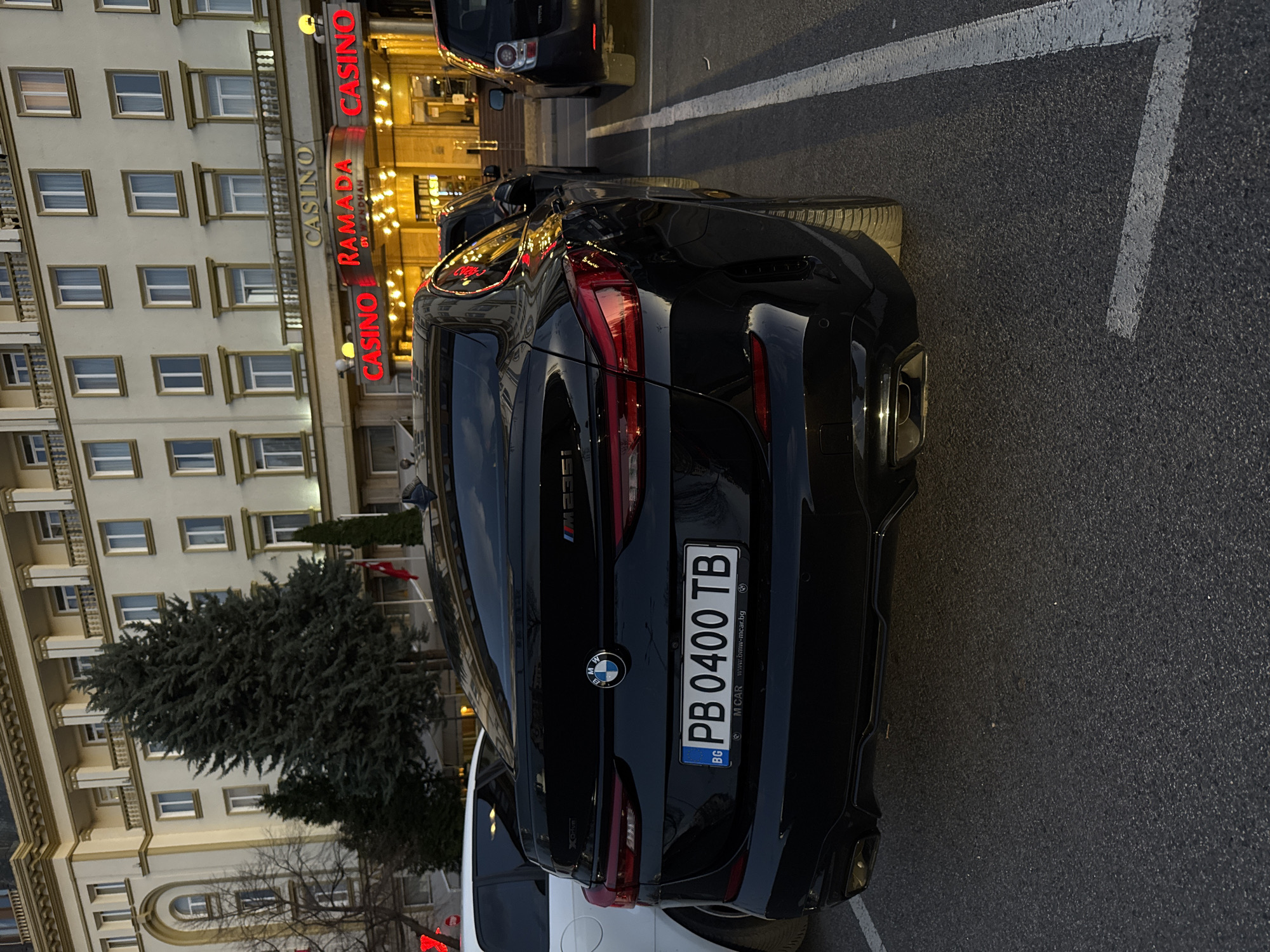 A dark-colored BMW SUV with Bulgarian license plate "PB 0400 TB" is parked in front of the Ramada by Wyndham Plovdiv Trimontium Hotel, identified by its large "CASINO RAMADA" sign. The hotel's exterior is characterized by numerous windows and decorative molding. A coniferous tree is to the left of the car. The parking surface is dark asphalt with painted white lines. Another car, possibly a white SUV, is parked partially visible behind the BMW. A dark-colored Toyota is parked farther ahead.