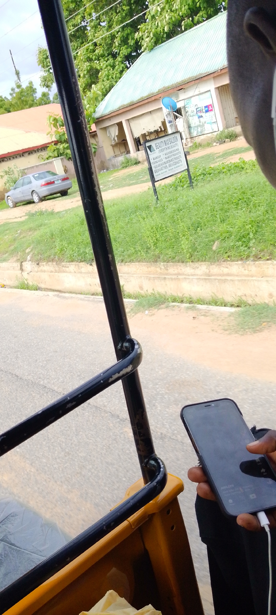 The image captures a street scene from the perspective of someone riding in a commercial tricycle (Keke Napep) in Jalingo, Nigeria. The interior of the tricycle, visible in the foreground, consists of black metal support bars and a yellow frame, with a crumpled light-colored plastic bag on the seat. On the right, a person's right ear and right hand are visible, holding a black smartphone with a white charging cable plugged into its port. The phone screen is reflective, but faint interface elements, including "YouTube" and text like "AJELERI", are discernible.
Outside the vehicle, a grey asphalt road is visible in the mid-ground. Beyond the road is a green grass verge with sparse vegetation. In the background, two single-story buildings are prominent. The central building features a light green corrugated metal roof and light-colored walls, partially obscured by green trees. In front of this building, a black and white sign advertises "BEAUTY MAX SALOON (EVERYTHING BEAUTY)", listing services such as "MAKEUP", "HAIRDRESSING", "FIXING NAILS", "MAKEUP PRODUCTS", and "HAIR AND HAIR MAINTENANCE PRODUCTS". Two Nigerian phone numbers, 08104467909 and 08133384379, are printed at the bottom of the sign. A small blue satellite dish is affixed to the side of this building. To the left, another building with an orange roof is visible, with a silver sedan car parked on a dirt patch in front of it. Overhead, power lines stretch across the frame against a bright, overcast sky.