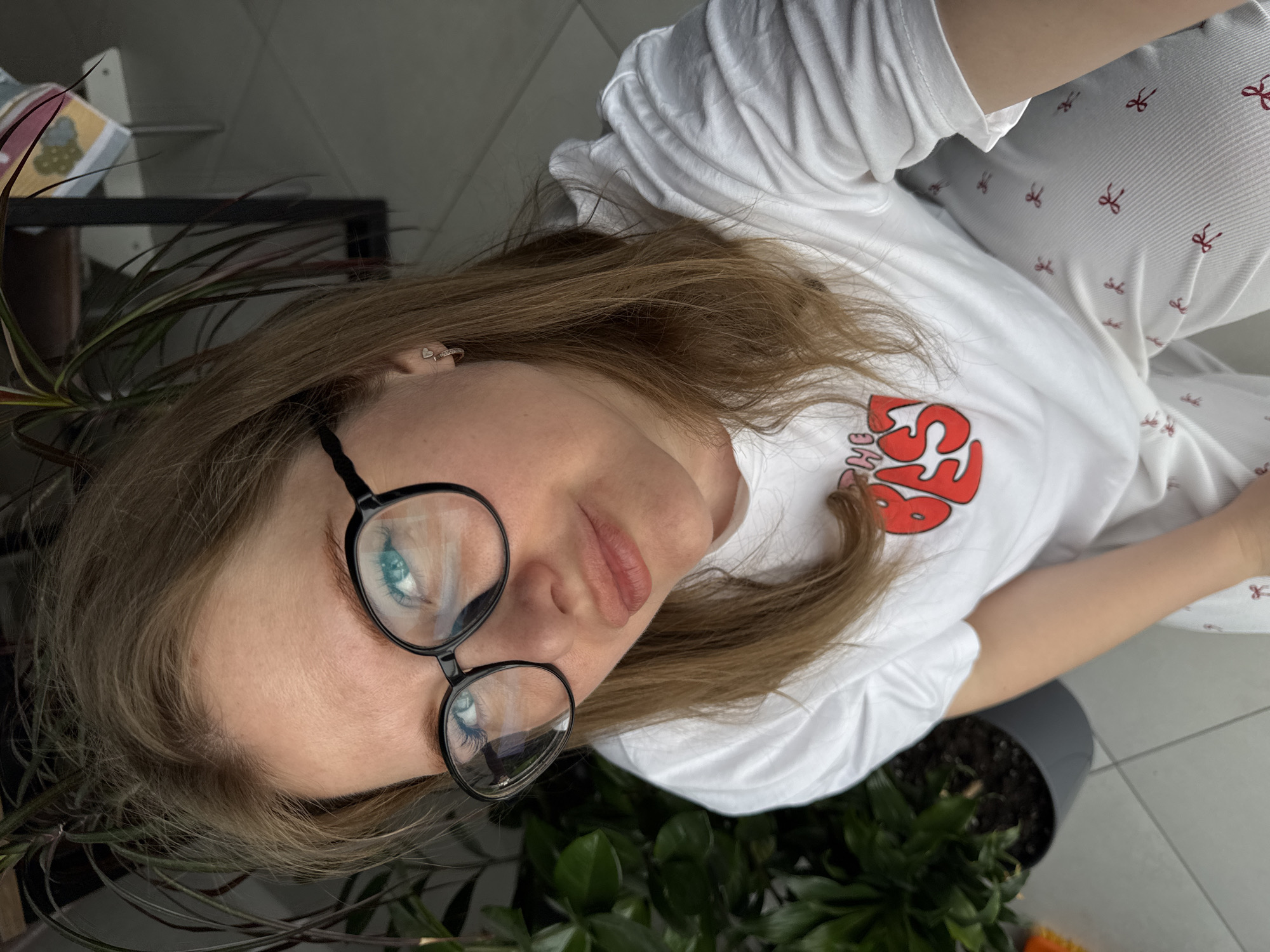 A woman with fair skin and light brown hair is wearing black-framed glasses, a white t-shirt with a red graphic, and white pants patterned with red bows. She is positioned among indoor plants in a gray planter. The location is likely in Горный, Russia.