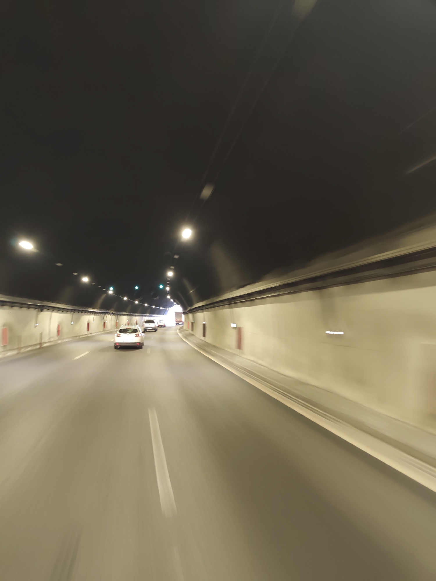 The image shows a view from inside a tunnel, likely a highway tunnel given the presence of multiple lanes and vehicles. The tunnel has a dark, arched ceiling with numerous lights illuminating the interior. The walls of the tunnel are a light, neutral color, with some recessed lighting and what appear to be red emergency call boxes visible.
Several vehicles are present on the road, moving away from the viewer and towards the light at the end of the tunnel. The closest vehicle is a white SUV or hatchback, followed by other vehicles further ahead, including what appears to be a white passenger car and a truck. The motion blur indicates that the image was taken while in motion, likely from a moving vehicle. The road surface is asphalt with visible lane markings.
The context provided about the location, Gorno Kamartsi, Bulgaria, suggests this is a public infrastructure scene. The overall impression is one of travel through an enclosed, man-made passage.