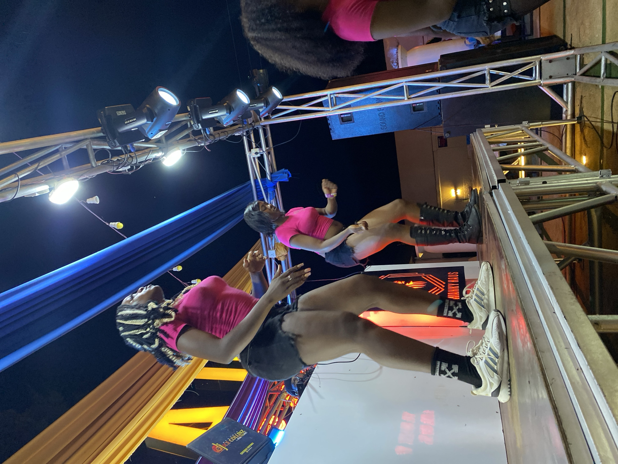 In Gura Top, Nigeria, at an outdoor nighttime event, three women are performing on a raised stage. The woman on the left, wearing a pink crop top, black distressed shorts, black socks with white stripes, and white athletic shoes, is captured mid-dance with her arms extended. To her right, another woman in a pink crop top and black distressed shorts is also dancing, her body turned to the side. A third woman, partially visible on the far right, wears a pink top and denim shorts. The stage is illuminated by stage lights and a large speaker labeled "SOUND PRO" is visible. The background features draped fabric in yellow and blue hues, and a dark night sky.