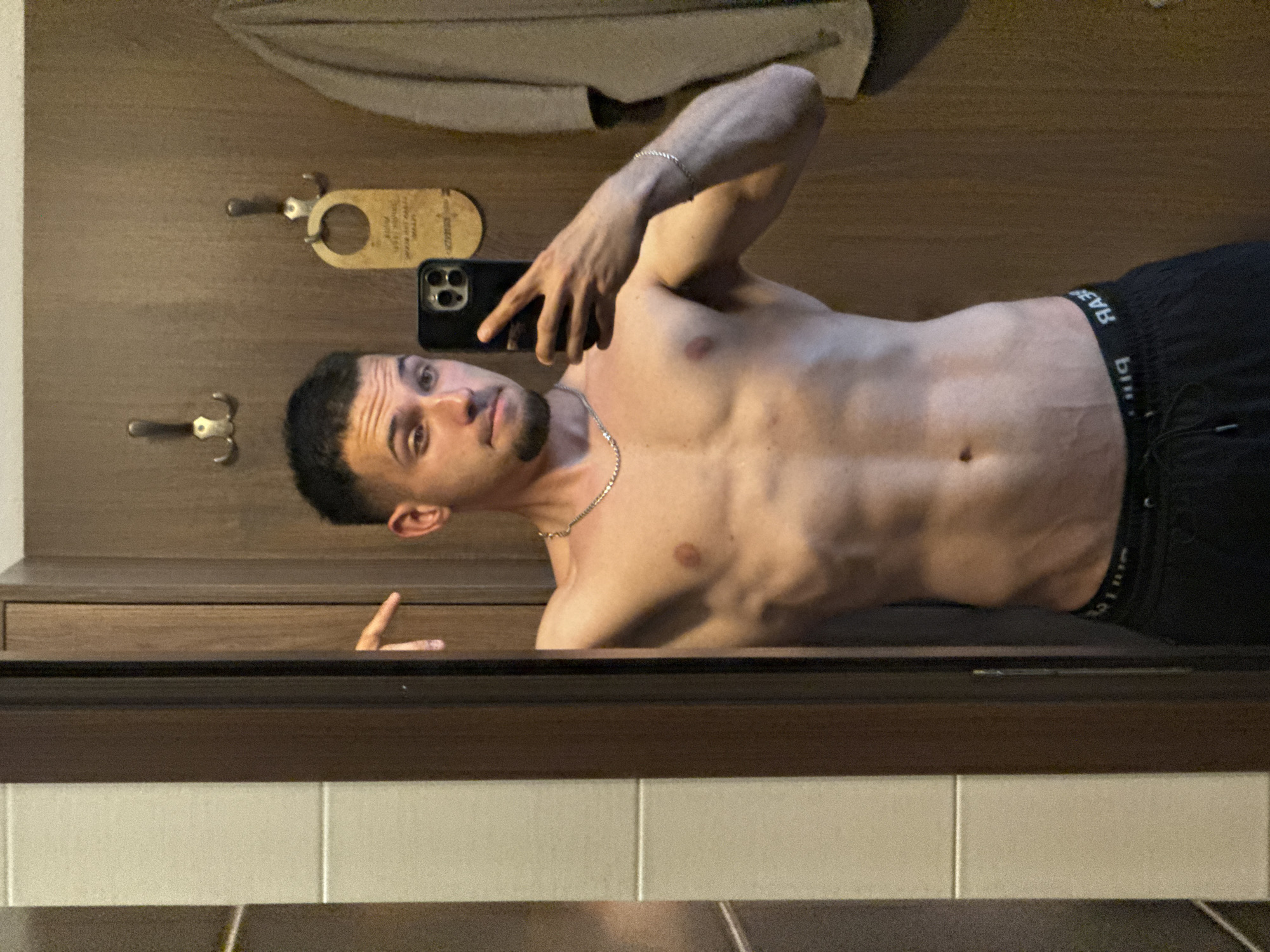 A shirtless man with dark hair and a goatee is taking a selfie in a mirror. He is wearing black shorts and a silver chain necklace. The background includes a wooden wall with hooks and a "do not disturb" sign, and tiled walls. The location cannot be determined to be Veliko Tarnovo, Bulgaria from the provided image.