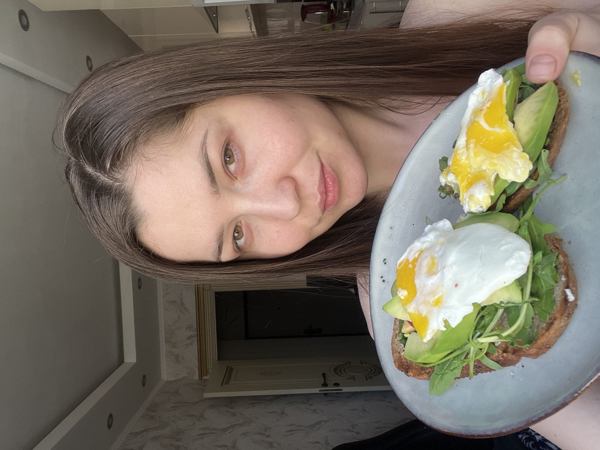 A young woman with long brown hair and fair skin is holding a plate with two pieces of toast topped with avocado, greens, and poached eggs. The picture appears to be taken indoors, possibly in a kitchen or dining area of a residence. The background shows elements of a wall and possibly a door with ornate trim. It is not possible to verify that the location is in the city of Khimki, Russia based on the image alone.