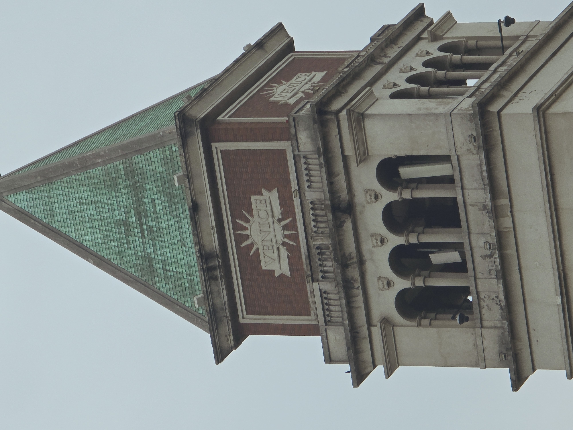 This is a close-up, eye-level, medium shot of the upper section of a clock tower. The tower is constructed of stone and brick, with a green tiled pyramidal roof. The facade features architectural details such as arches, columns, decorative stone faces, and a balcony. Two prominent rectangular panels on the brick section of the tower bear the word "VENICE" in raised lettering, each surrounded by a sunburst motif. A small black bird is perched on the edge of the balcony on the left side of the tower. A security camera is attached to the right side of the tower, mounted on an arm extending from the building. The background is a solid, overcast sky. The image does not depict the city of Xiaoshan District, China; it is a depiction of a tower in Venice.