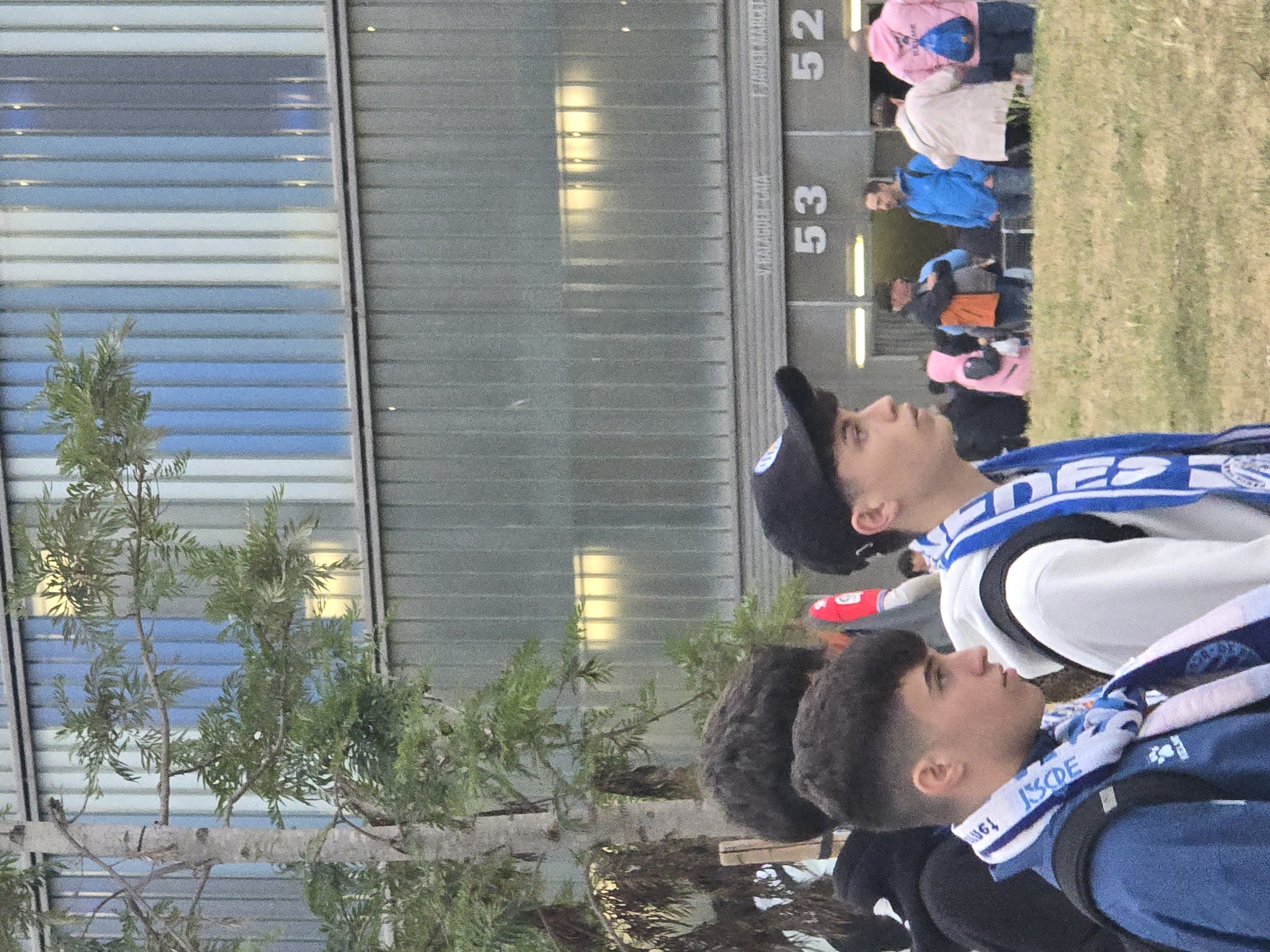 The image shows a group of people near a building, likely a stadium, in Cornellà de Llobregat, Spain. Two young men are prominently featured in the foreground wearing team scarves. Behind them, individuals are seen queued near numbered entryways (53, 52). The background features a modern building façade, and a small tree is visible on the left. The presence of team scarves and the numbered entryways suggest the scene is near a sports venue before or after an event.