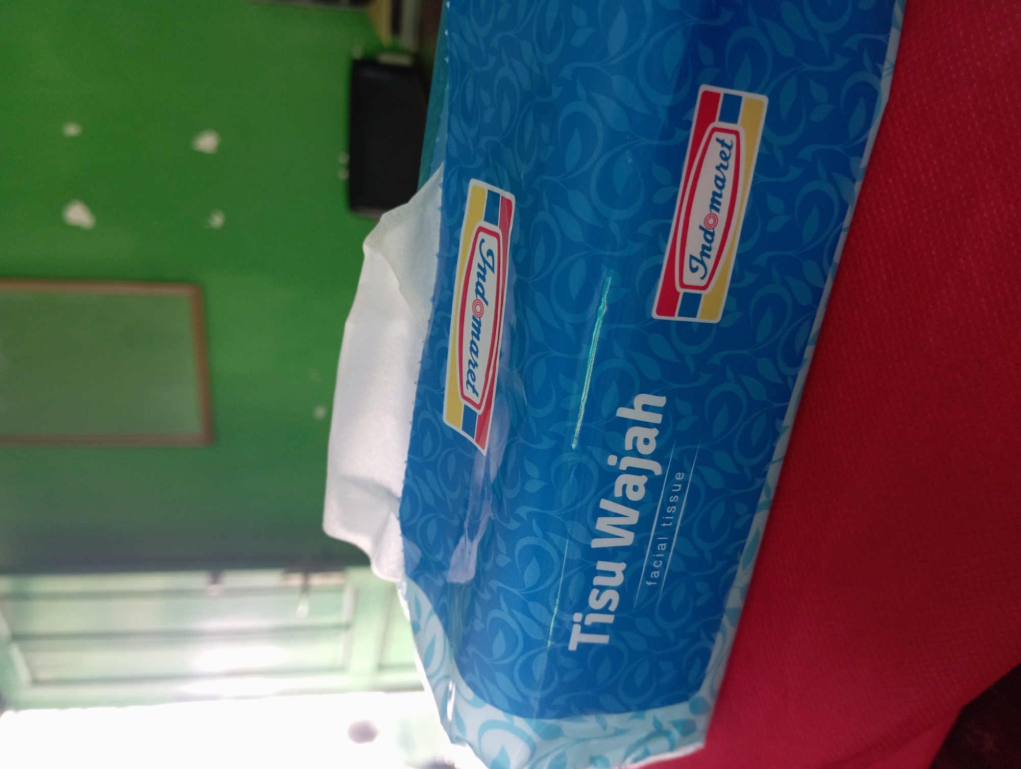A tissue box with the brand name "Indomaret" is shown in close-up, resting on a red surface. The tissue box is blue with a leafy pattern and has the words "Tisu Wajah" and "facial tissue" printed on it. The background is blurred, showing a green wall and a door. The scene appears to be indoors, possibly in a residential or commercial setting in Tegalciut, Indonesia.