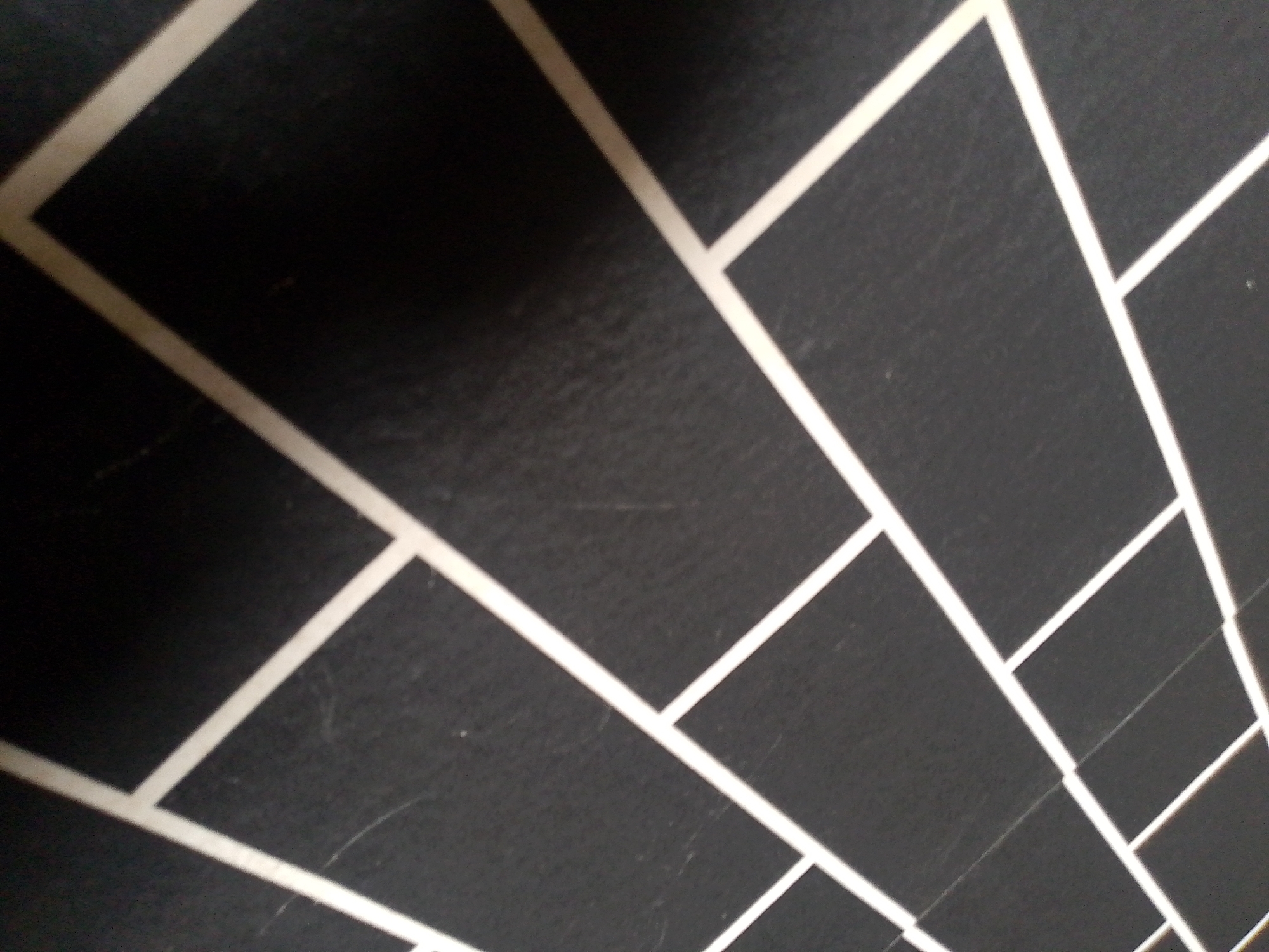 The image shows a close-up, abstract view of a dark gray surface with a pattern of thin, light-colored lines. The lines are arranged in a grid-like, perspective pattern, suggesting they might be part of a wall, floor, or some decorative element. The lighting creates shadows and highlights, emphasizing the texture of the dark surface, which appears slightly matte and possibly textured. There are no discernible people, objects, or actions visible in the image. The scene is static and devoid of any contextual clues that would allow for identification of a specific location within Jauro Yinu, Nigeria, or any other city.
