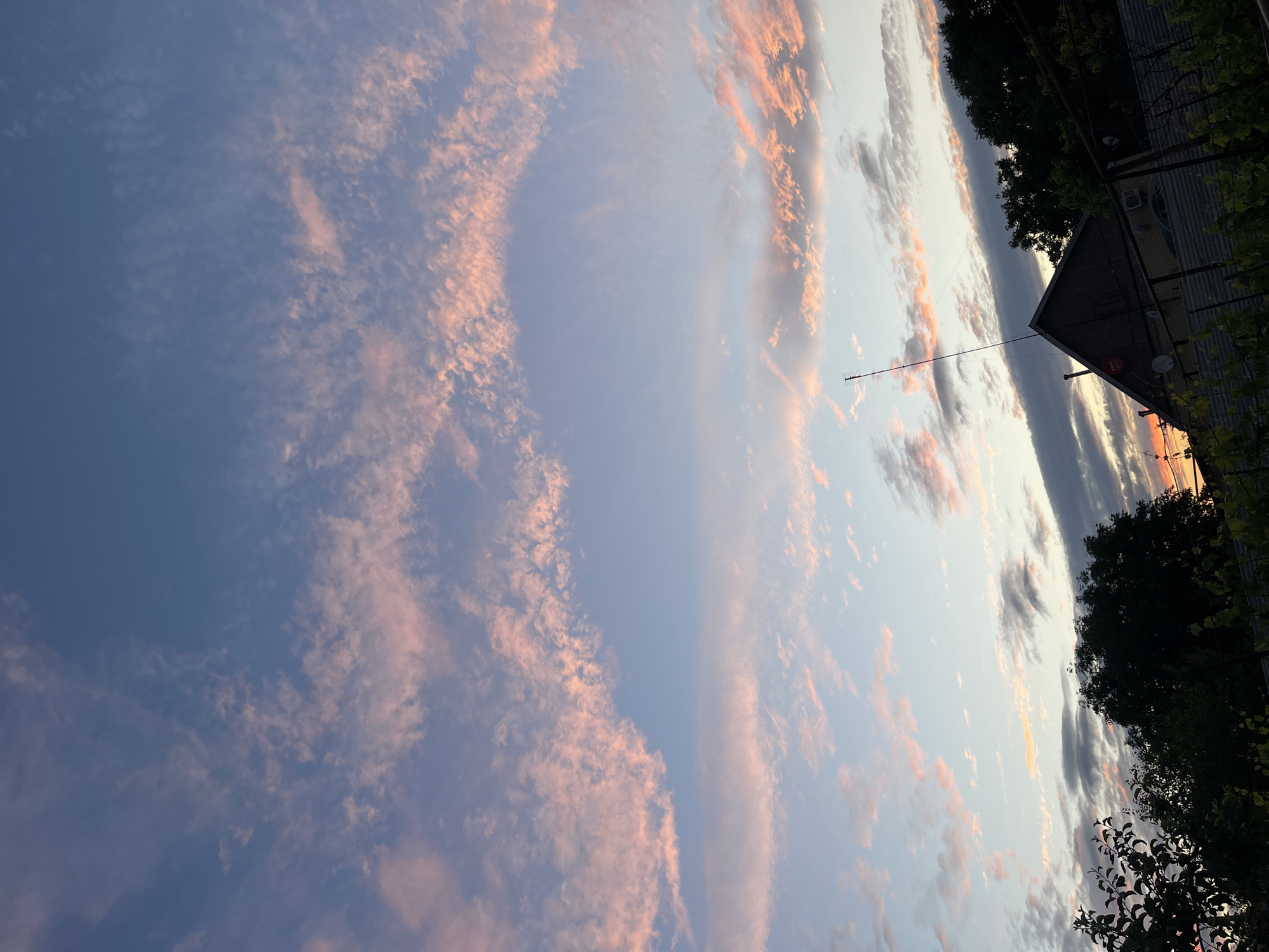 This is a daytime photograph, taken outdoors, likely during sunset, showcasing a sky filled with wispy clouds tinged with pink and orange hues against a gradient of blue. In the lower portion of the image, silhouetted against the sky, are the dark shapes of trees, a fence, and the roof of a building. A tall antenna extends upwards from the roof. The overall scene depicts a tranquil, possibly rural or suburban, landscape. The provided information states the location is the city of Sredny, Russia, however, there are no specific identifying landmarks or characteristics within the image that confirm this location.