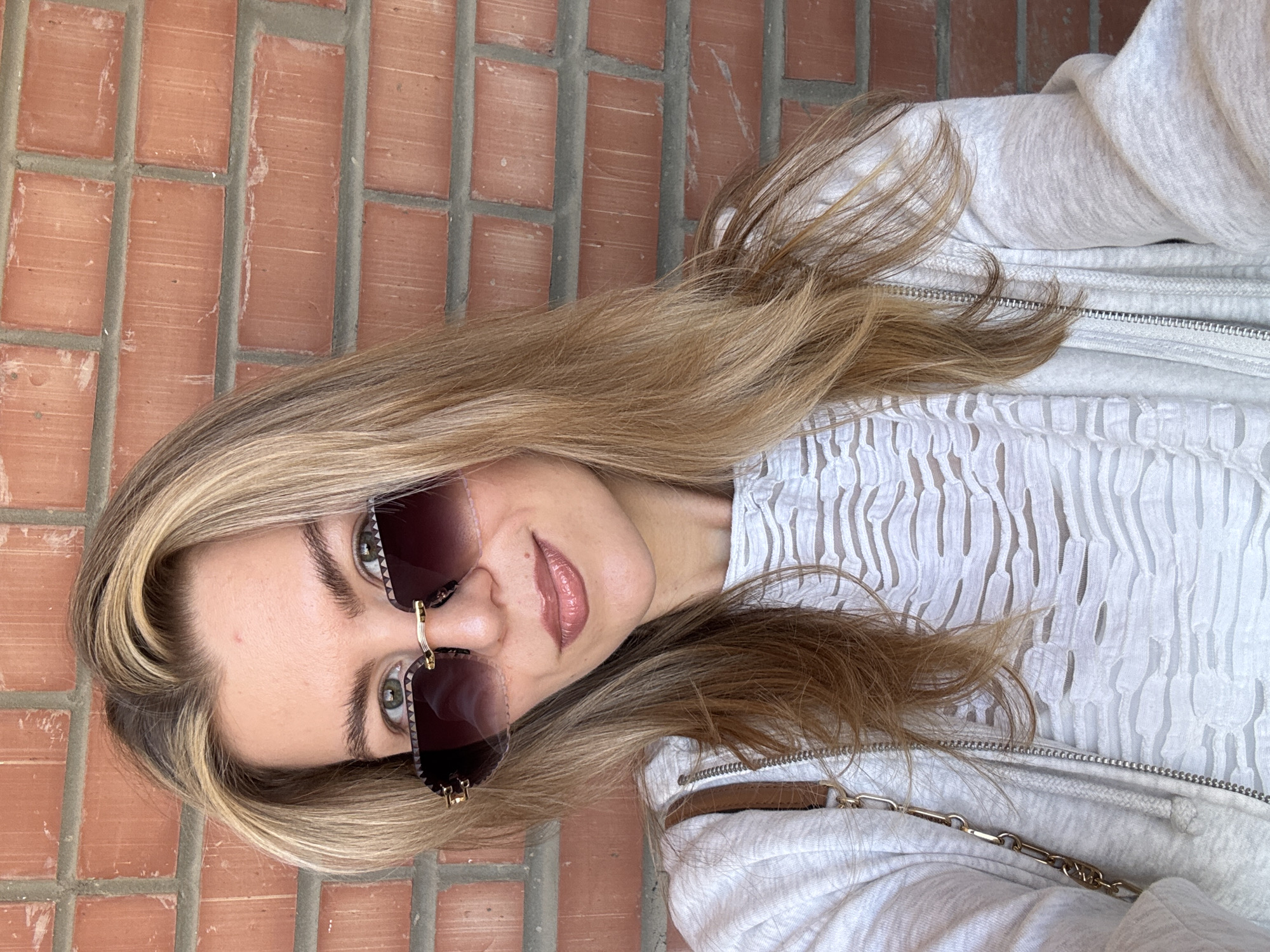 A blonde woman wearing sunglasses and a light-colored jacket is positioned against a brick wall. The background suggests an urban setting, though the specific location within the city of Unknown, Unknown is not discernible. The woman is the primary subject, and her interaction with the environment is limited to her positioning against the wall. The scene is static, capturing a moment in time.