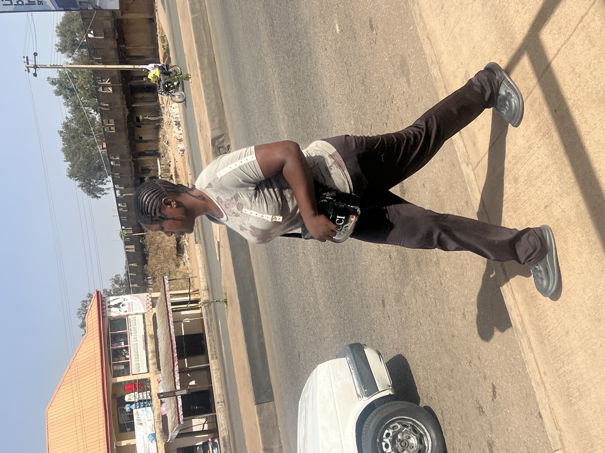 The image captures an outdoor street scene in Jos, Nigeria, under bright daylight with strong shadows indicating a sunny sky. The primary subject is a person, likely an adult, standing or walking on a paved sidewalk next to a road. They have dark skin and braided hair, and are wearing a light grey short-sleeved t-shirt with a subtle floral or abstract pattern, dark brown or black track pants, and grey slip-on shoes (resembling Crocs). They are carrying a small black bag with white text, which appears to say "GUCCI" on its side.
In the mid-ground, a white car's front bumper and wheel are partially visible, suggesting it is parked or moving slowly. Beyond the road, there are buildings and other structures. To the left, a commercial building with a reddish-brown roof and multiple signs is visible. One prominent sign reads "GOOD FABRICS ENTERPRISE" and includes a phone number "07033285735". Above it, another sign shows text including "DANDY" and "PLAYWORLD", with "24HRS" also partially visible on another sign. Further down the street, there's a multi-story concrete building that appears to be unfinished or disused, along with a wooden utility pole carrying electrical wires. A person on a motorcycle, possibly carrying green leafy produce, is visible riding away in the distance near the utility pole. The background also features some trees and what looks like a dirt or unkempt area. The clear sky is a pale blue.