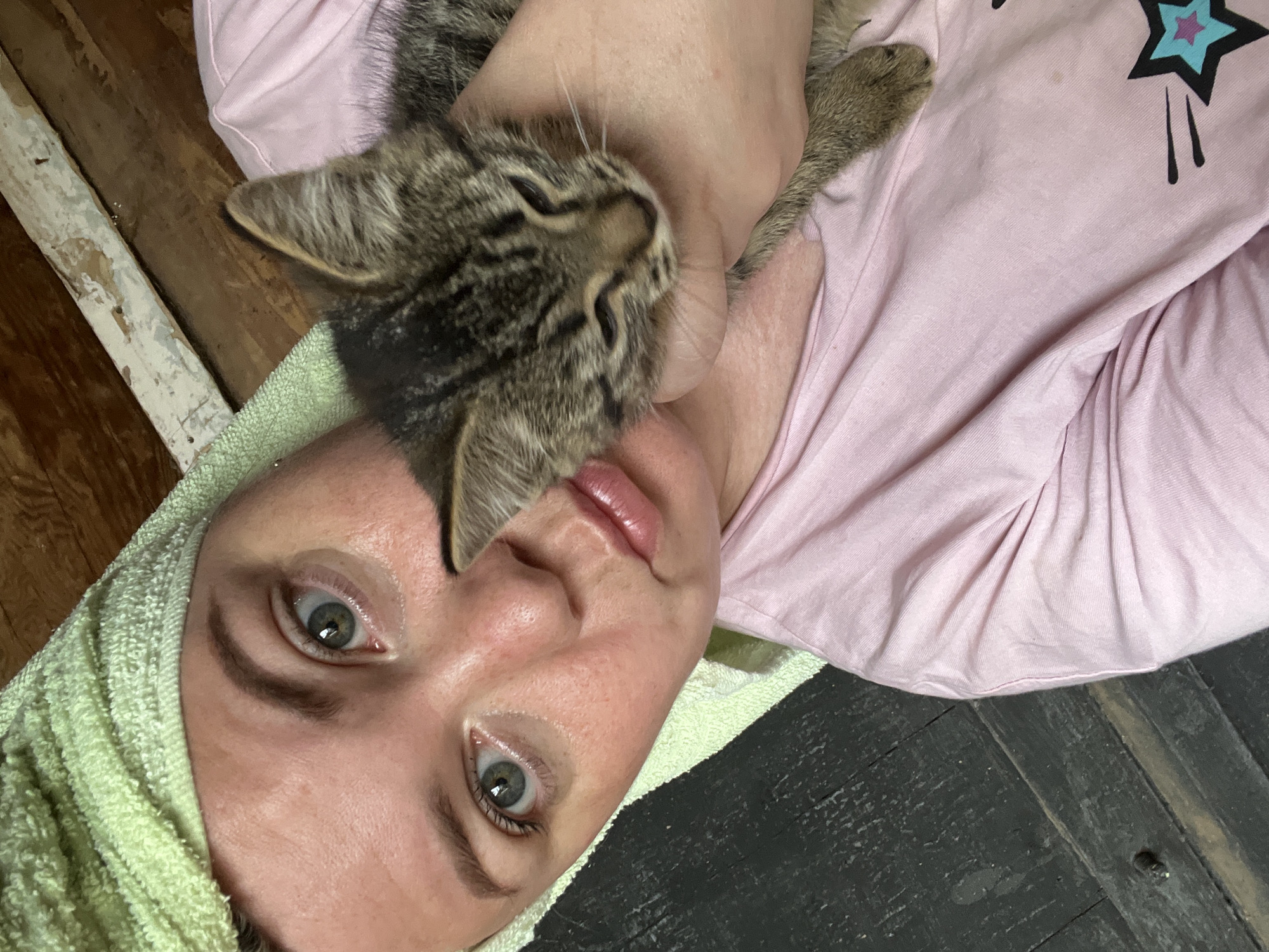 A close-up, selfie-style photo shows a person with a light green towel wrapped around their head, holding a small tabby kitten. The person is wearing a pink t-shirt with a star graphic. Their eyes are open and looking at the camera. The kitten is held close to the person's face, with its eyes partially closed. The background appears to be a wooden interior, possibly a rustic or unfinished room. The lighting is natural and comes from the front. The image does not contain enough contextual information to determine its location within the city of Fokino, Russia.