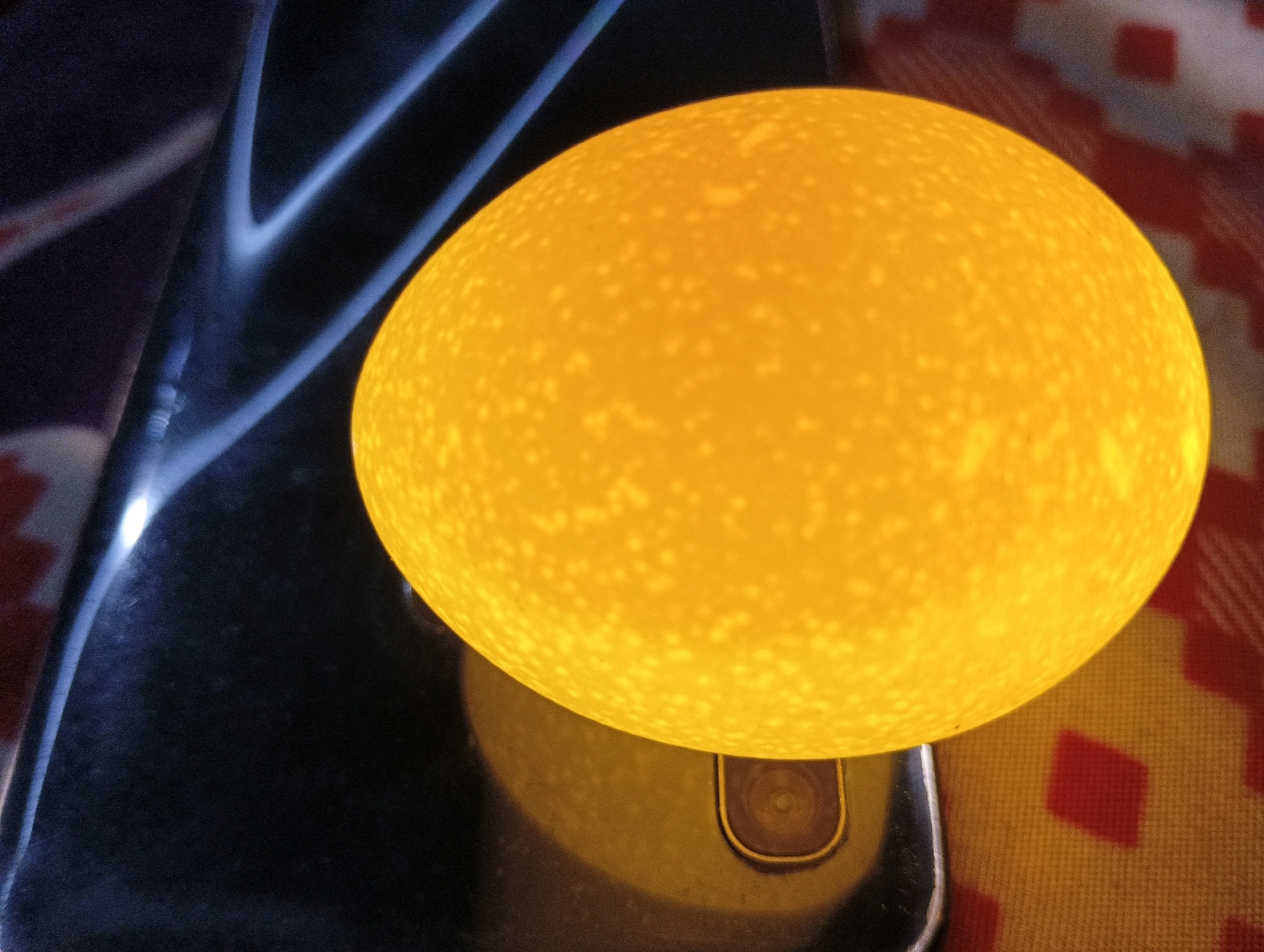 An egg, illuminated from within and displaying numerous small, bright spots on its surface, rests on a dark, reflective object with curved blue highlights. The object appears to be a smartphone, as evidenced by a visible camera lens on its surface. The egg is positioned on the right side of the frame, partially obscuring the smartphone. To the right of the egg, a textured surface with a red and yellow diamond pattern is visible. The overall scene is captured from a high angle, with shallow depth of field. The lighting suggests a controlled environment, possibly for examination or display. There is no indication of the city of Muzaffarnagar, India, or any specific environmental context beyond the immediate objects and background.