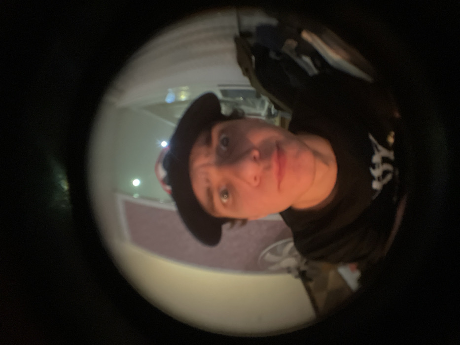 Through a fisheye lens, a person wearing a dark hat and a black t-shirt with white lettering looks directly at the camera. The background is blurry, but a hallway with a door and flooring is visible, lit by several lights. A fan is also somewhat discernible behind the person. The lens's distortion creates a wide, rounded view with a dark, black frame surrounding the central image.
