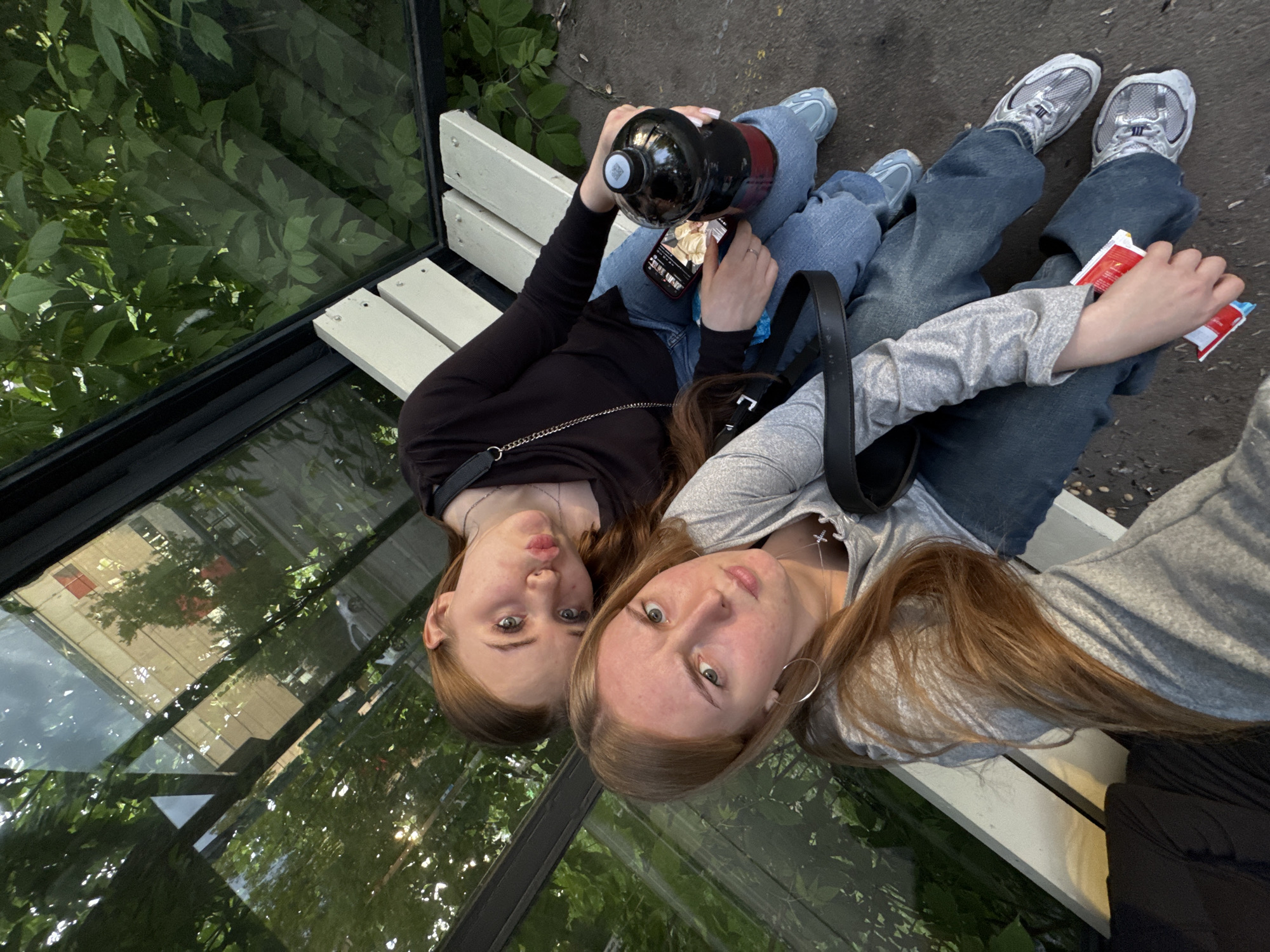 Two young women are seated on a white wooden slatted bench inside a glass-paneled public transport shelter in Perm, Russia. The shelter features dark framing, and its glass panels reflect a green tree canopy, a street with a white vehicle, and a multi-story building with a red-framed window or facade element on an upper floor. The woman in the foreground, positioned to the left, has long light brown hair and is wearing a grey long-sleeved shirt, blue jeans, and grey and white sneakers. She holds a red and white packaged item in her left hand. She wears a silver hoop earring in her left ear and a silver cross pendant on a necklace. Her gaze is directed towards the camera with a serious expression. The second woman, positioned behind and to the right, has long reddish-brown hair and is wearing a black long-sleeved top, blue jeans, and light blue sneakers. She has light pink painted fingernails. She rests her head on the first woman's left shoulder and holds a dark-colored plastic bottle containing a reddish liquid in her right hand, and a smartphone displaying an image of food in her left hand. Her lips are puckered in a pouting expression directed towards the camera. The ground outside the shelter is dark asphalt or concrete.