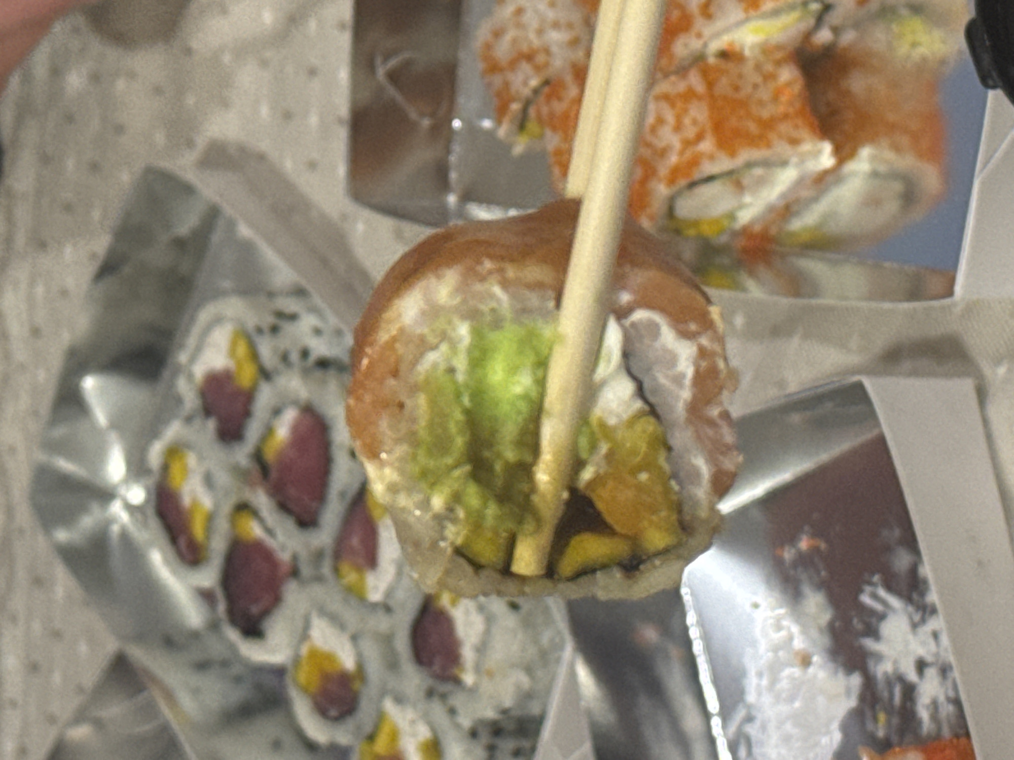 The image displays a close-up of various sushi rolls in takeout containers, captured in an indoor setting. The central subject is a single sushi roll held by light-colored wooden chopsticks, poised for consumption. This particular roll features a light brown outer layer, which could be salmon or seared tuna, encasing a filling that includes bright green avocado, creamy white sauce (likely cream cheese), and yellow or orange components, possibly mango or another vegetable.
In the background, two other types of sushi are visible within their disposable containers. To the upper right, several rolls are topped generously with vibrant orange roe, revealing white rice and green or yellow ingredients beneath. To the lower left, another container holds rolls characterized by white rice and black sesame seeds on the exterior, with fillings of distinct dark red, suggestive of tuna, alongside yellow-orange elements.
The scene appears to be a casual dining environment, possibly a home, given the takeout packaging. The background is a soft, light-colored, and slightly patterned surface. No text is visible in the image. This meal originates from Krasnoyarsk, Russia.