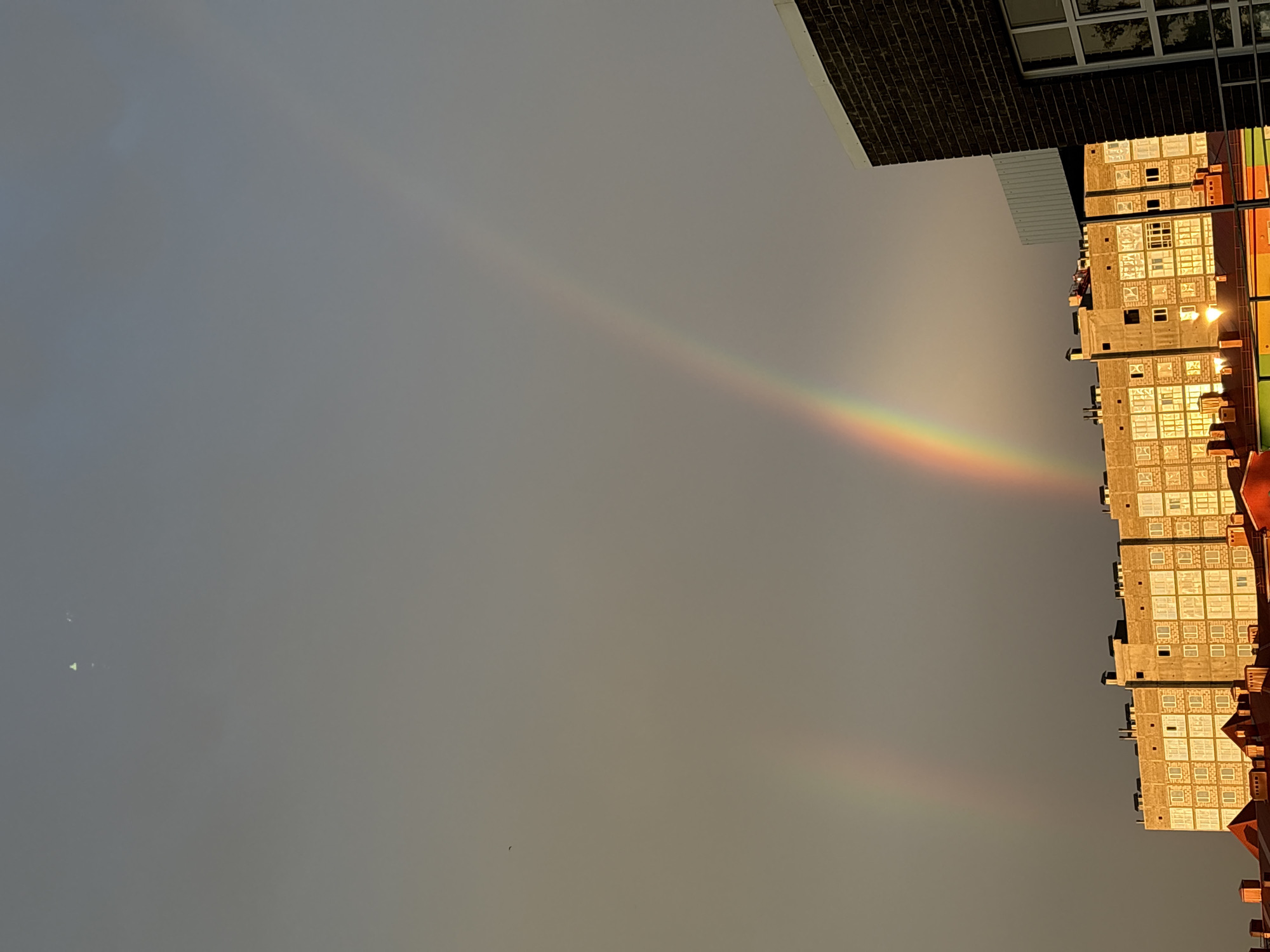 In Mikhaylovsk, Russia, a double rainbow arcs across a grey, overcast sky above a cityscape. The primary rainbow is prominent, displaying a vibrant spectrum of colors, while a fainter secondary rainbow is visible to its left. In the foreground, several buildings are partially visible. The largest is a multi-story apartment building with numerous windows, appearing to be under construction, as some sections are bare concrete. To the right, a darker brick building with a large window is present. Below the apartment building, the roofs of other structures, some with a reddish hue, are visible. The sky is illuminated by what appears to be the setting or rising sun, casting a warm glow on the buildings. There are no people or animals clearly visible in the scene. The overall atmosphere is one of a post-rain or stormy weather condition in an urban setting.
