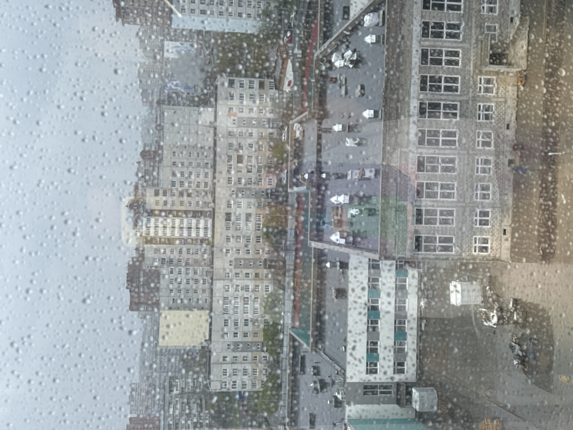 The image shows an urban landscape through a raindrop-covered window. The view includes multiple high-rise residential buildings with repetitive window patterns, suggesting an apartment complex or a densely populated urban area. Between the buildings, there are roads and parking areas. The environment appears to be overcast, as the sky is gray and the lighting is subdued. Rain obscures the view, creating a blurred and textured effect on the windowpane. There is no indication of people or specific actions taking place. The location is unknown, as there are no recognizable landmarks or signs.