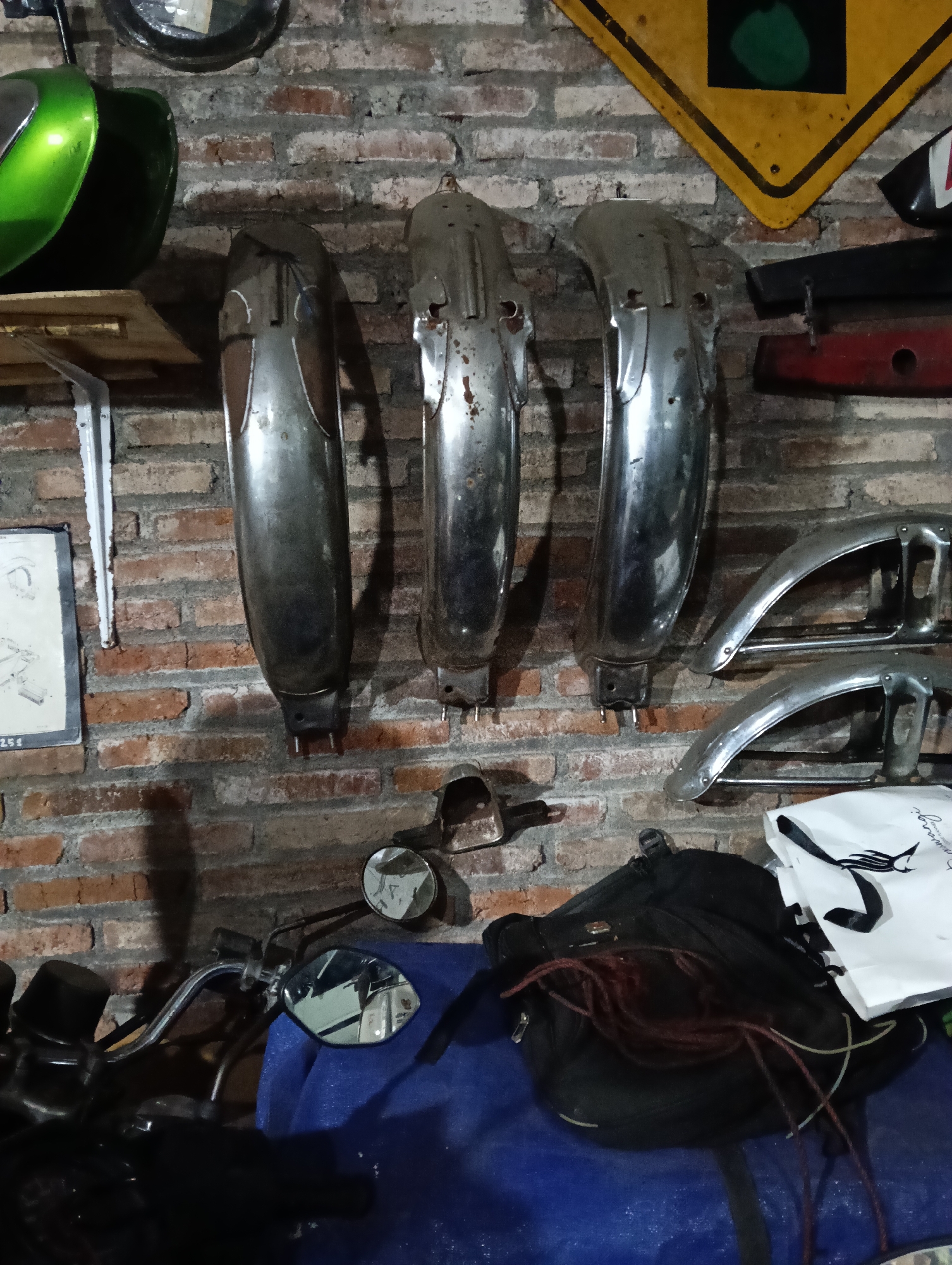 The image depicts a cluttered garage or workshop space, likely in Magelang, Indonesia. The backdrop is a rustic brick wall. Several vintage motorcycle parts are prominently displayed, including three large, curved metal fenders hanging vertically, two smaller chrome fenders mounted horizontally, and a green motorcycle fuel tank on a shelf. Other objects visible include a yellow road sign with a black and green circular motif, a black motorcycle bag with red cords, a white shopping bag with a logo, and parts of a motorcycle, including handlebars and mirrors. The scene suggests a collection of motorcycle parts or a repair area. There are no people visible in the image.