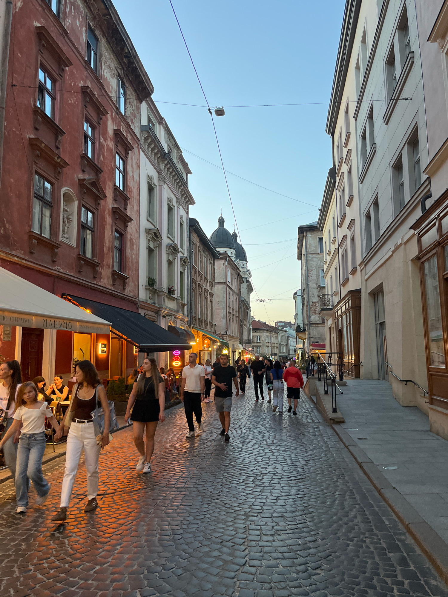 The image depicts a street scene in Lviv, Ukraine, likely during the evening, as evidenced by the warm lighting emanating from storefronts and street lamps. The cobblestone street is lined with historic buildings, characteristic of Lviv's Old Town. On the left, a building with a reddish-brown facade displays architectural details such as decorative window frames and what appears to be a niche with a statue. Below, an outdoor seating area for a cafe or restaurant is set up with umbrellas.
A diverse group of people are walking along the street, some appearing to be tourists or locals enjoying an evening stroll. They are dressed in casual attire. On the right side, a cream-colored building with multiple windows and a distinct entrance is visible, suggesting shops or residences. In the distance, church domes or cupolas can be seen, further confirming a European city setting, likely within Lviv's historical center. The sky is a clear gradient of blue and orange, indicating either dawn or dusk. The overall atmosphere is lively and characteristic of a popular urban thoroughfare.