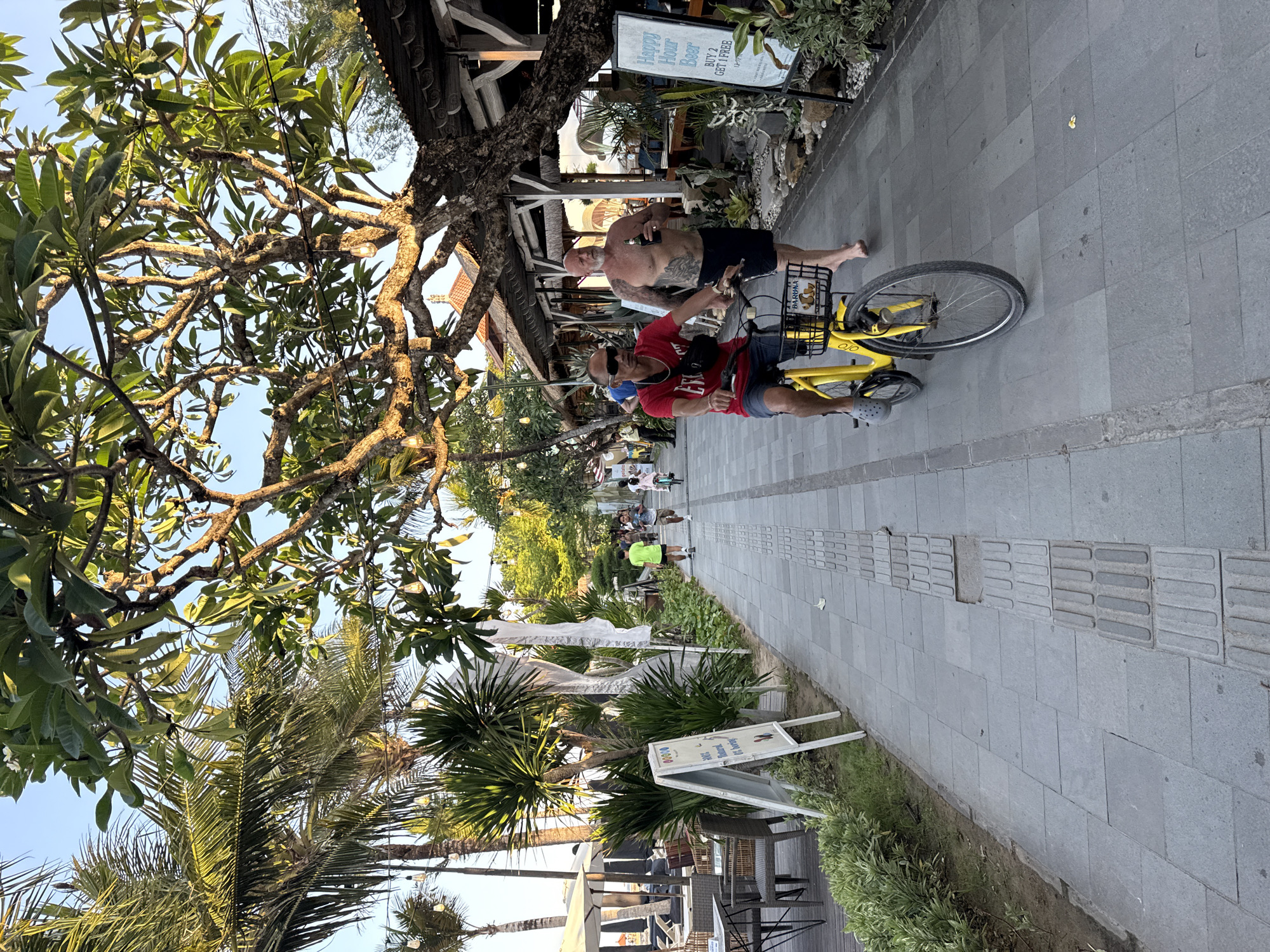 The image depicts a pedestrian walkway in the city of Denpasar, Indonesia, likely along a beach or tourist area, given the presence of palm trees, tropical foliage, outdoor seating, and a "Happy Hour Beer" sign. The walkway is paved with grey stone tiles, and a tactile paving strip runs along one side.
In the foreground, a man wearing a red t-shirt, sunglasses, and greyCrocs is riding a yellow bicycle. He has a black fanny pack slung across his chest. Behind him, to his right, a shirtless man with tattoos on his arms and chest is walking, holding a bottle in his right hand.
Further down the walkway, other individuals are visible: a man in a bright green shirt is jogging, and a child on a bicycle is riding ahead of him. A young child on a bicycle is also seen further in the distance, along with other pedestrians.
On the left side of the walkway, there are outdoor seating areas with tables and chairs, reminiscent of a cafe or restaurant. Umbrellas provide shade over some of the seating. A white A-frame sign stands on the left, displaying some colorful circles and text.
Lush green vegetation and mature trees, including palm trees and a large tree with a thick, gnarled trunk, line the walkway. String lights are visible hanging from the trees. Buildings with thatched roofs and white structures are present in the background, suggesting commercial establishments. The sky is clear and blue, indicating daylight. The overall scene suggests a lively and sunny day in a tropical environment.