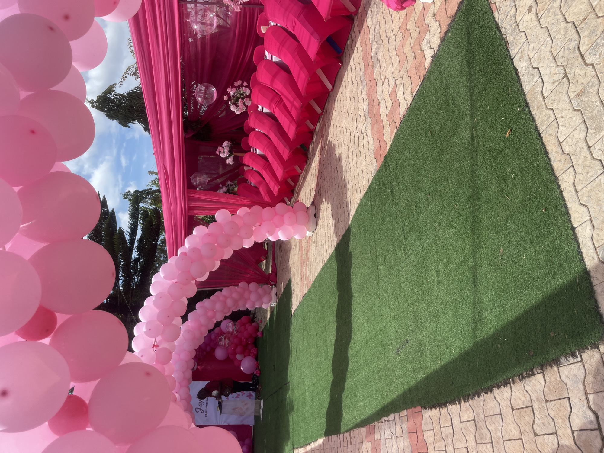 An outdoor event space in Gura Top, Nigeria, is decorated with an abundance of pink balloons and fabric drapes. A pathway covered in green artificial turf leads towards a seating area with chairs covered in pink fabric. Behind the seating area, a pink tent-like structure is adorned with additional pink balloons and floral arrangements. A banner with the word "Joy" is visible on the left side of the image. The ground surrounding the turf path is paved with light-colored bricks. The sky above is partly cloudy.