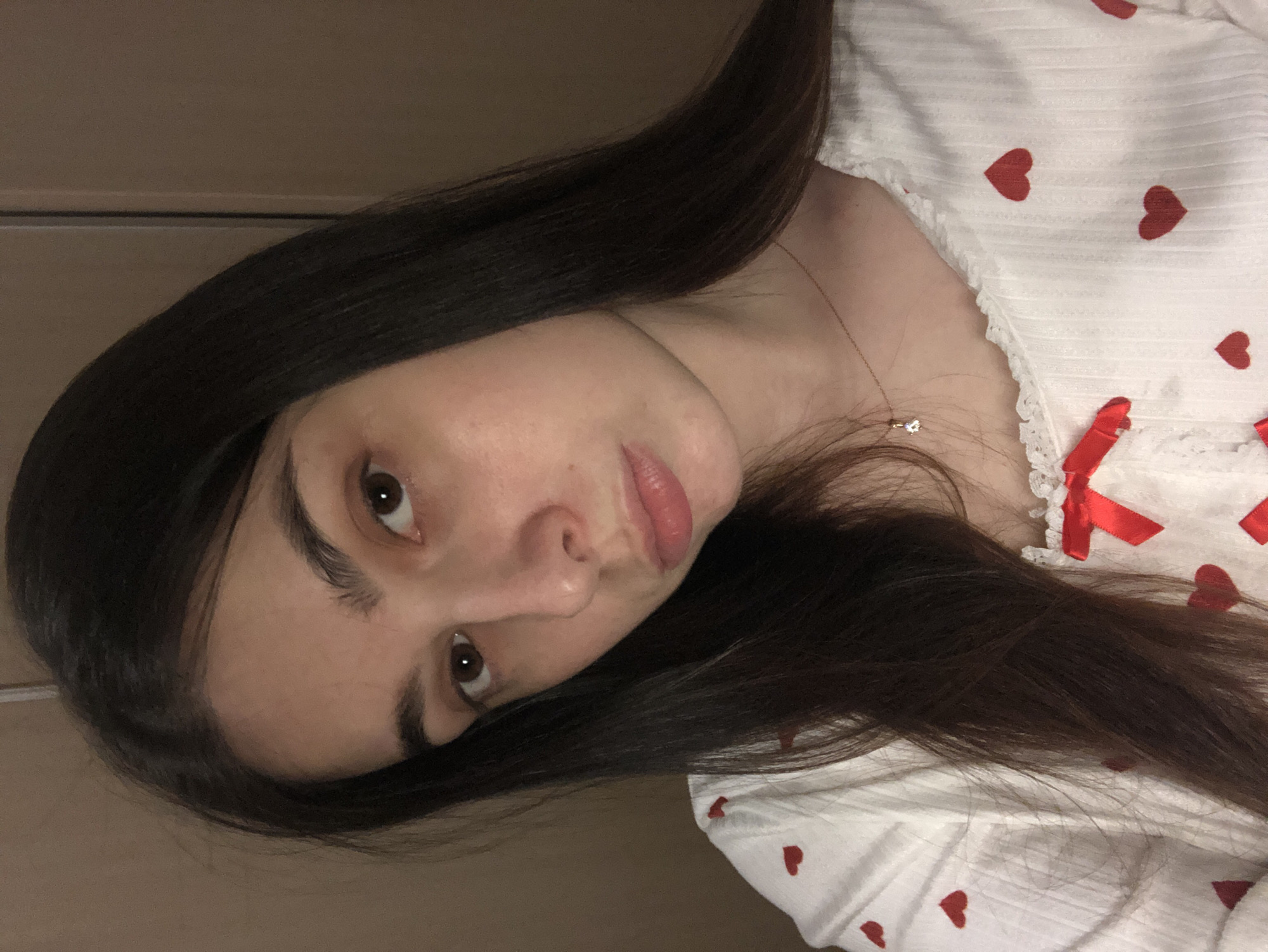 A woman with long, dark brown hair is photographed indoors, wearing a white garment adorned with red heart patterns and a small red bow. She is positioned at an angle, with her head tilted to the side. Her gaze is directed towards the camera. A gold necklace is visible around her neck. The background appears to be a plain wall. There is no visual information to infer the location to be in Noviy, Russia.
