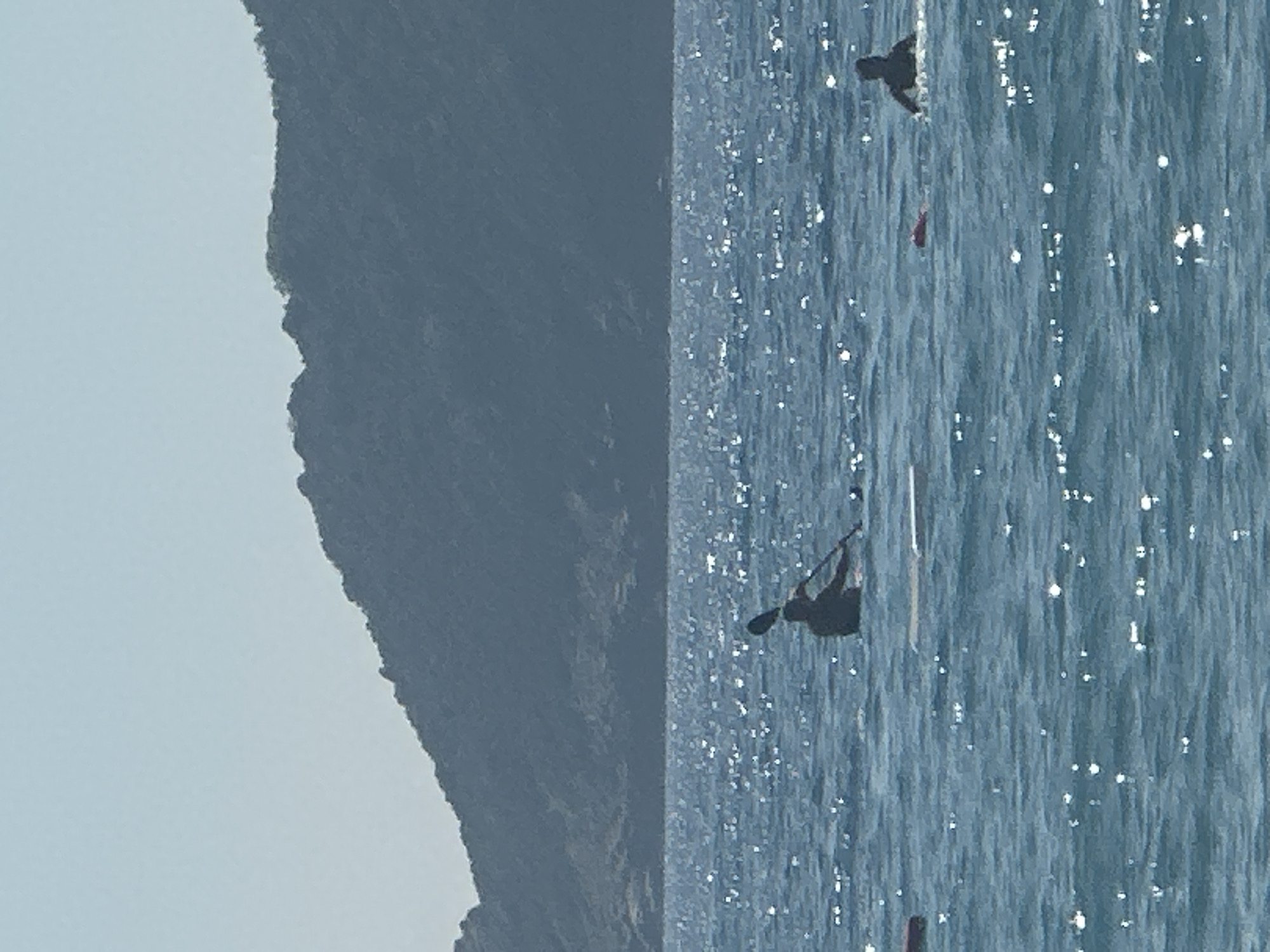 In Boreti, Montenegro, two individuals are engaged in water activities on a sunny day. In the foreground, a person is seated in a kayak, holding a paddle. To the right and further back, another individual is partially submerged in the water, appearing to swim or float. In the distance, a mountainous coastline is visible, with the sea reflecting the sunlight. The scene suggests a leisurely outdoor recreational setting.